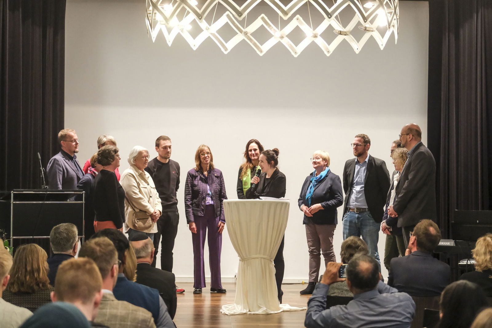 Mehrere Personen stehen vor einem sitzenden Publikum, eine Frau mit Mikrophon an einem weißen Stehtisch in der Mitte.