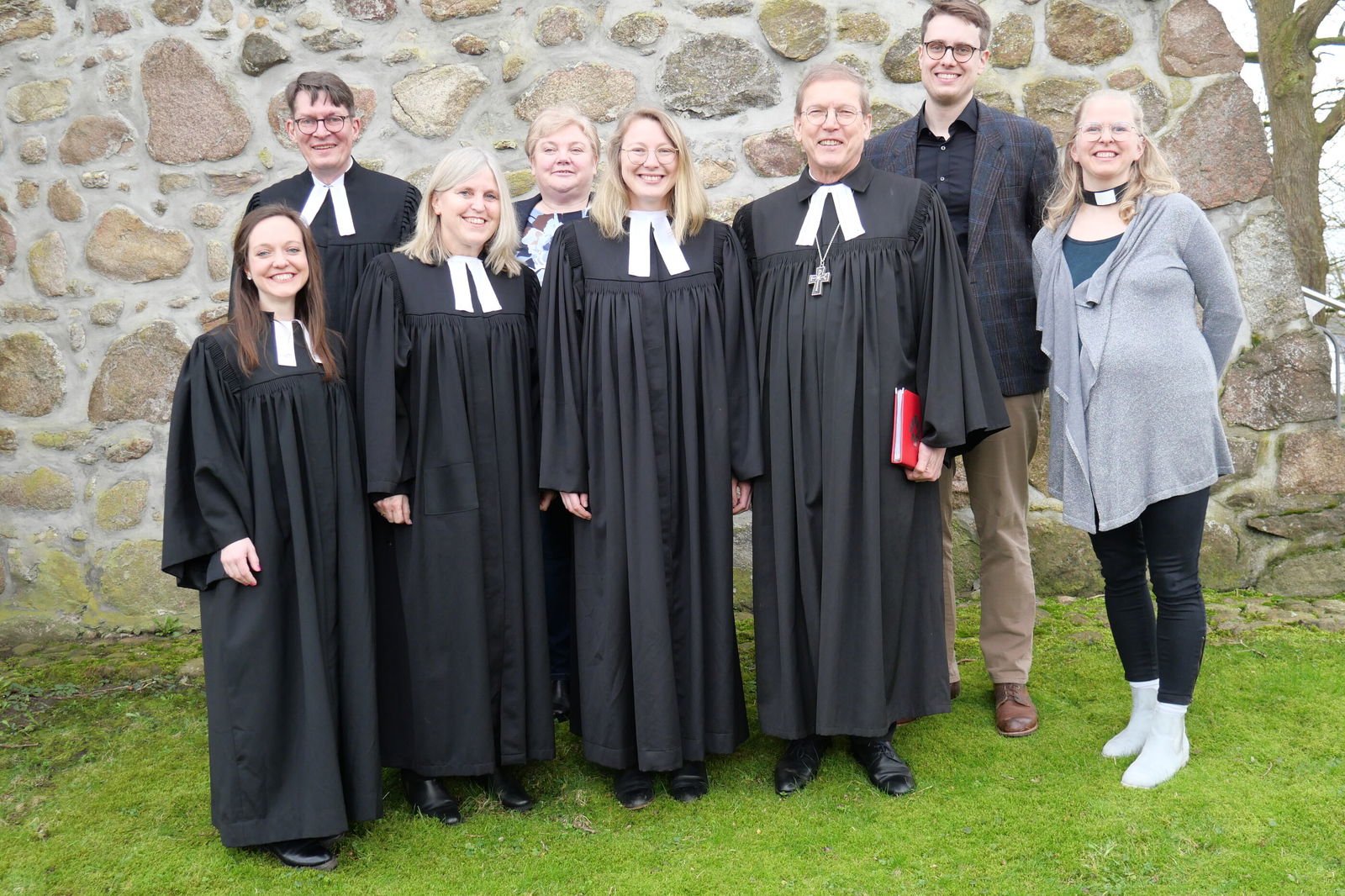 Eine Gruppe von menschen steht vor einer Kirchenmauer auf grünes Rasen. Bis auf zwei Personen tragen alle einen Tlar. Die beidne Personen erchts sind festlich gekleidet. Alle lächeln in die Kamera.