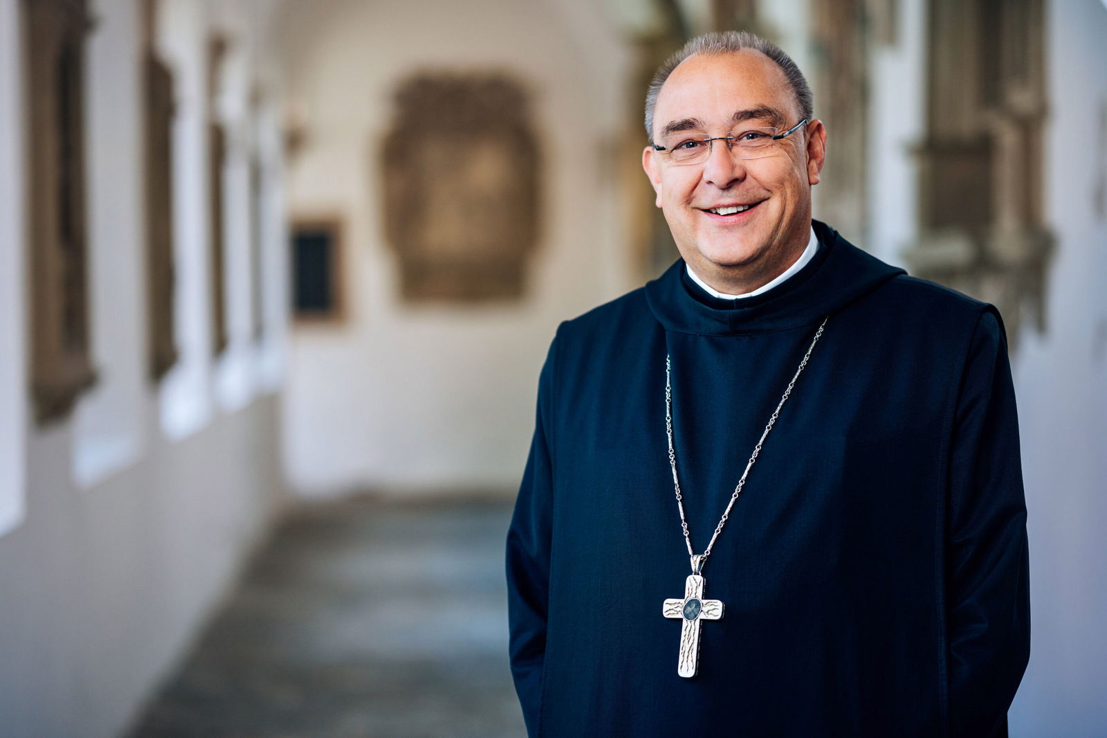 Eine als Mann lesbare Person steht im Bischofsornat in einem historischen Gang.