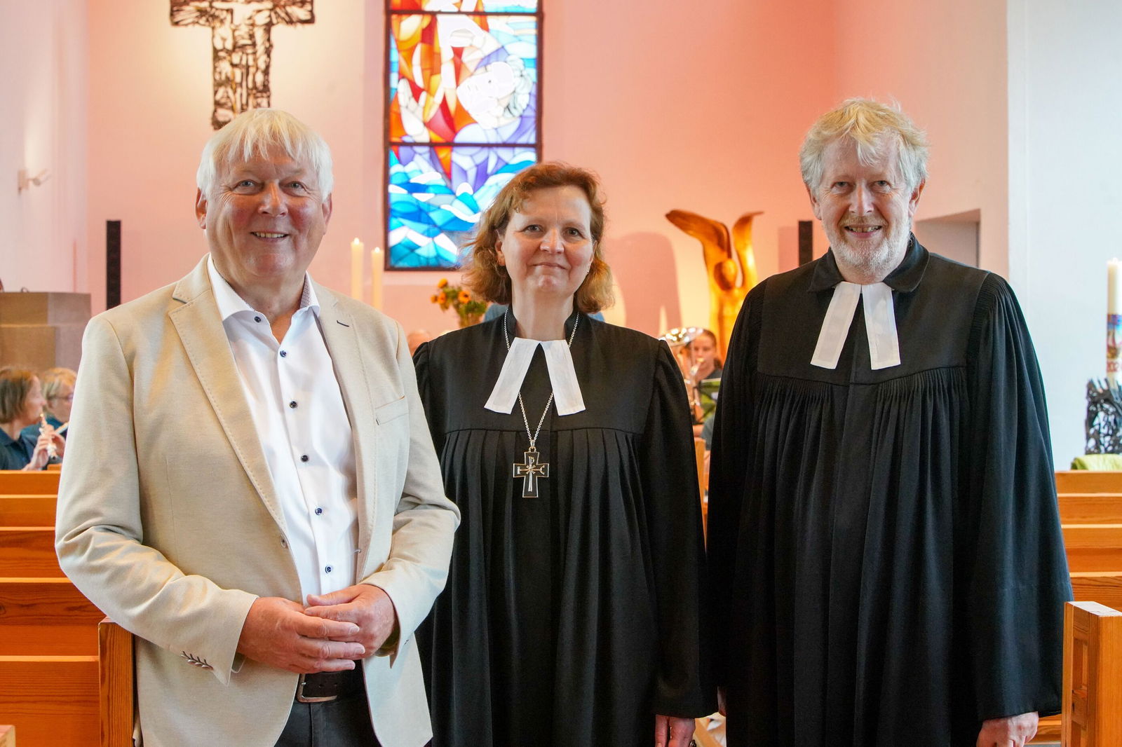 Ein Mann im einem weißem Anzug steht neben einem Mann und einer Frau im Talar in einer Kirche. Alle lächeln.