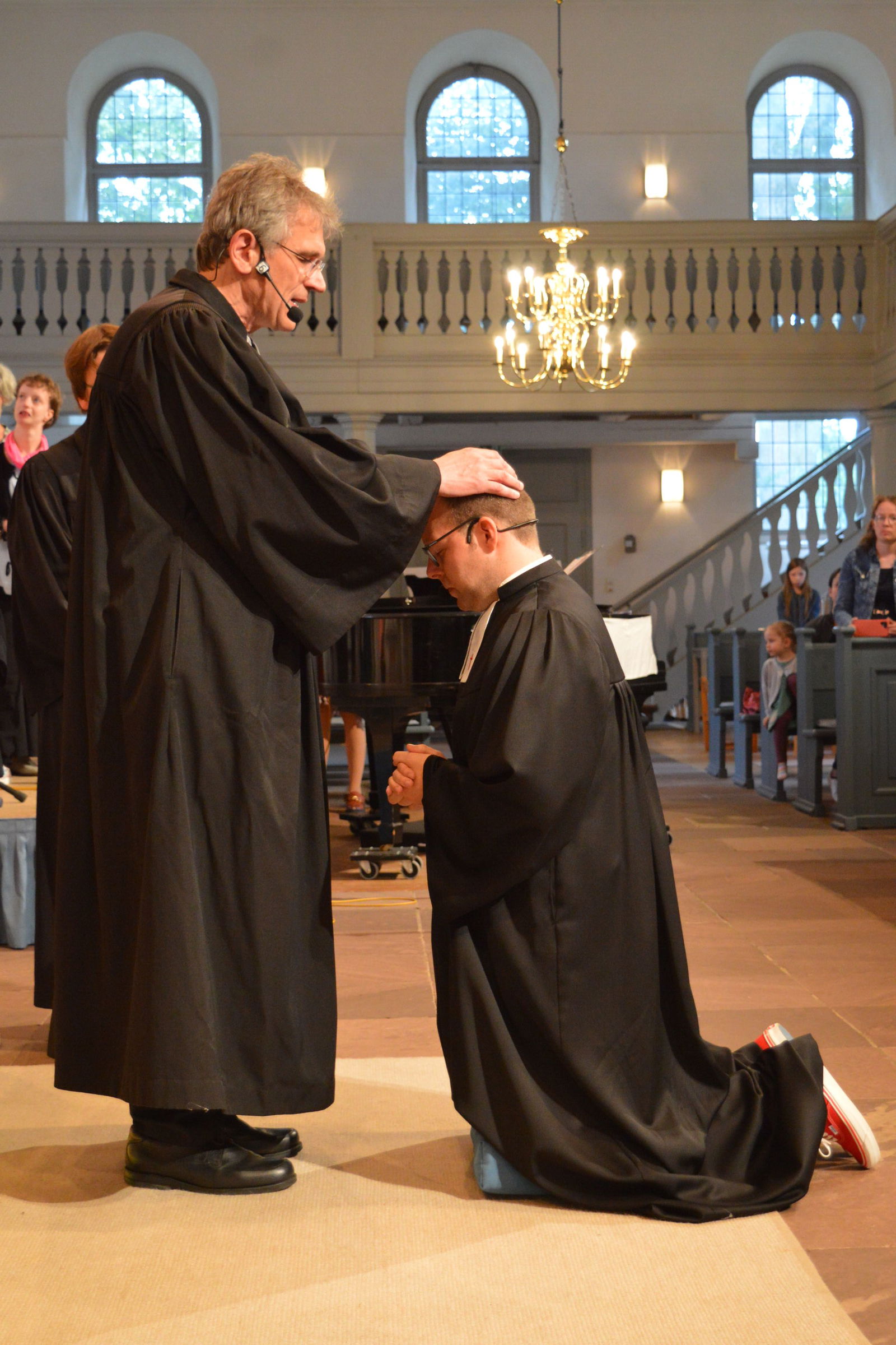Zwei Pastoren in Talar einer Kirche. Einer kniet. Er wird ordiniert.