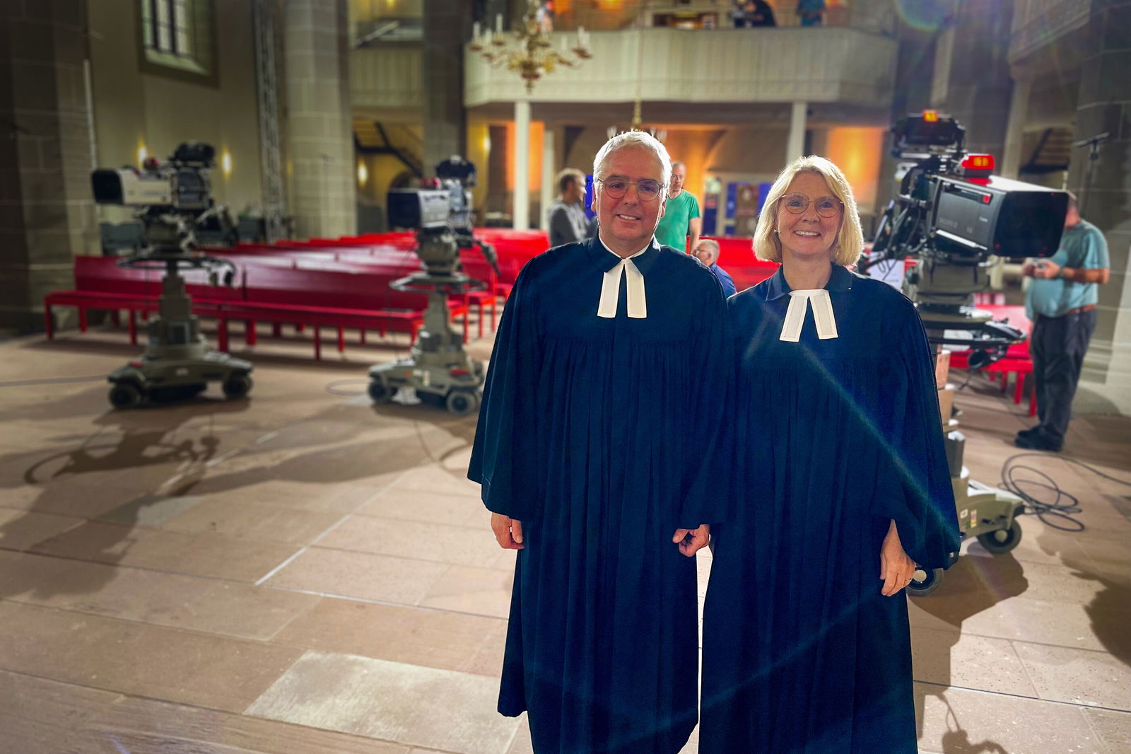 Eine männlich und eine weiblich gelesene Person, beide in Talaren, stehen in einer Kirche. Im Hintergrund einige Fernsehkameras.