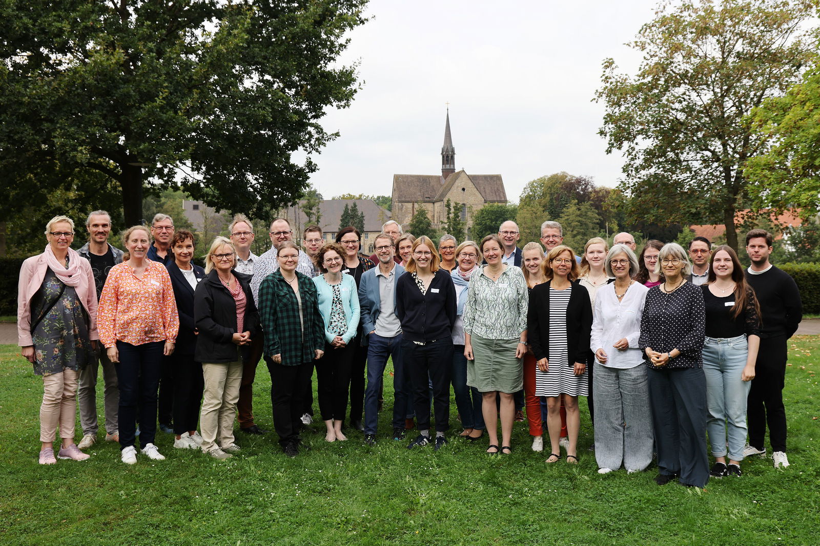 Eine größere Gruppe von als Männern und Frauen lesbaren Personen posiert auf einer Wiese mit einer Kirche im Hintergrund.