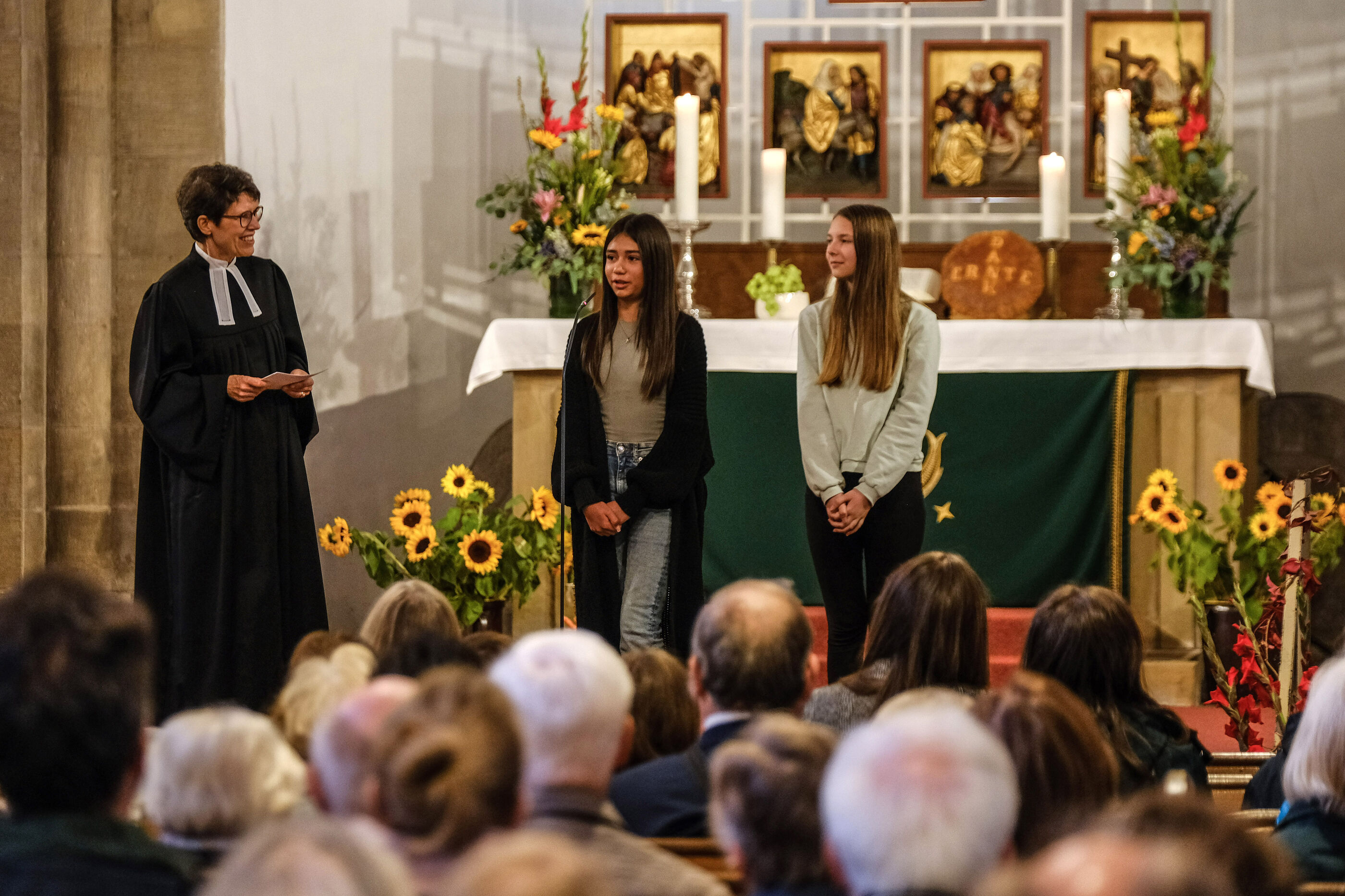 Zwei junge Menschen und eine ältere Person im Talar stehen in einer Kirche vor dem Altar, in den Bänken sind Menschen von hinten, sitzend, zu sehen. Den Altar schmücken Feldgaben, Sonnenblumen, Kürbisse und mehr.