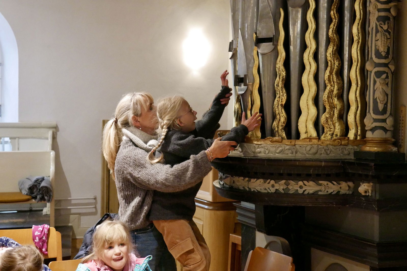 Eine erwachsene, weiblich gelesene Person hält ein Kind auf den Armen, das seine Hände zu großen silbernen Orgelpfeifen ausstreckt.