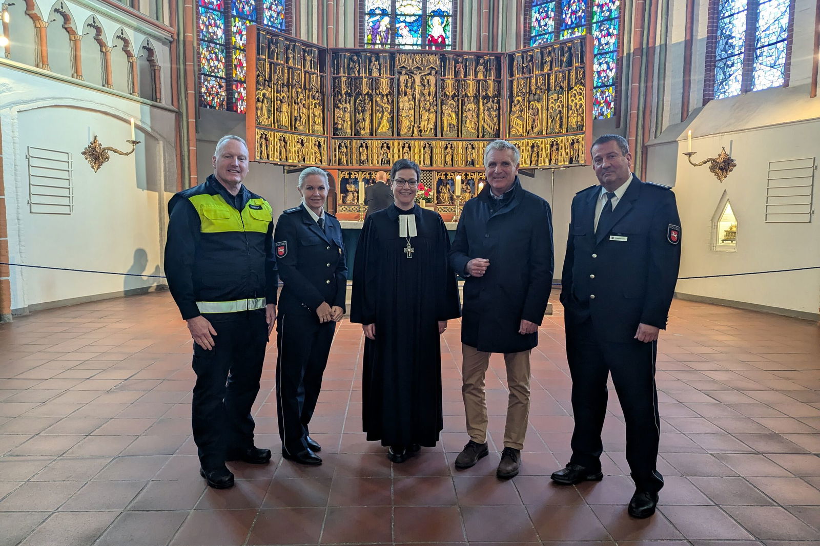 Fünf Personen, zwei weiblich und drei männlich gelesene stehen zusammen in einer Kirche. Einer trägt eine Einsatzjacke der Polizei, eine einen Talar, einer Feuerwehr-Uniform.