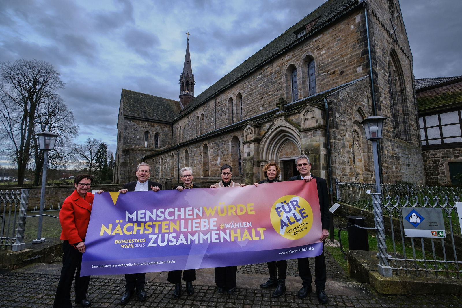 Vier weiblich und zwei männlich zu lesende Personen halten ein Banner. Im Hintergrund ist eine Kirche zu sehen.