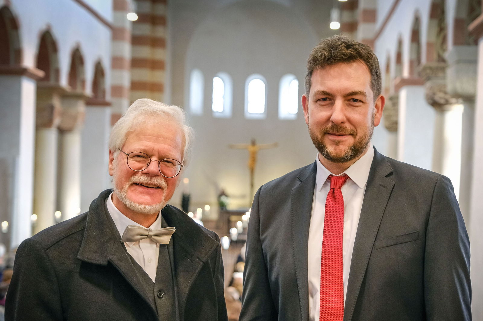 Zwei männlich gelesene Personen, eine älter mit weißem Haar und Brille und Fliege, neben einer jüngeren mit rotem Schlips in einer Kirche.