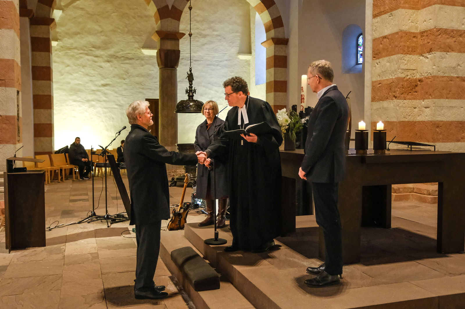 Drei Menschen stehen erhöht vor einer anderen Person. Die mittlere trägt Talar und reicht der entgegenstehenden Person die Hand.