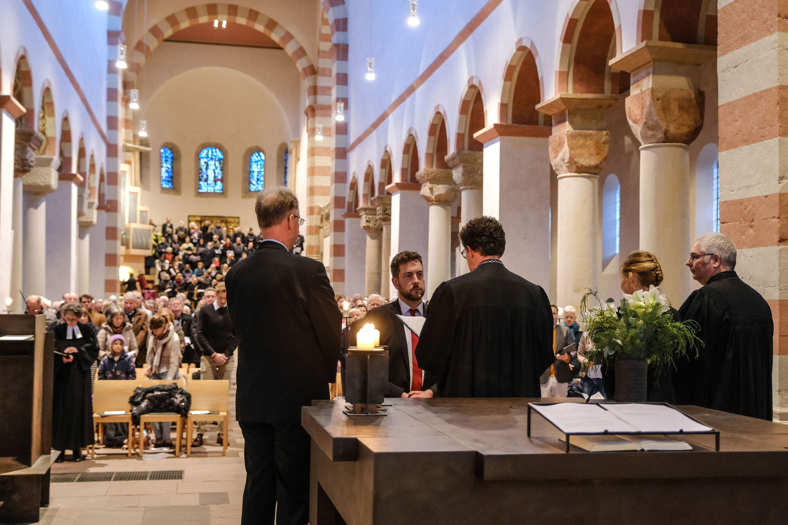 Blick in die Länge einer Kirche, die voll besetzt ist. Im Vordergrund stehen mehrere Personen.