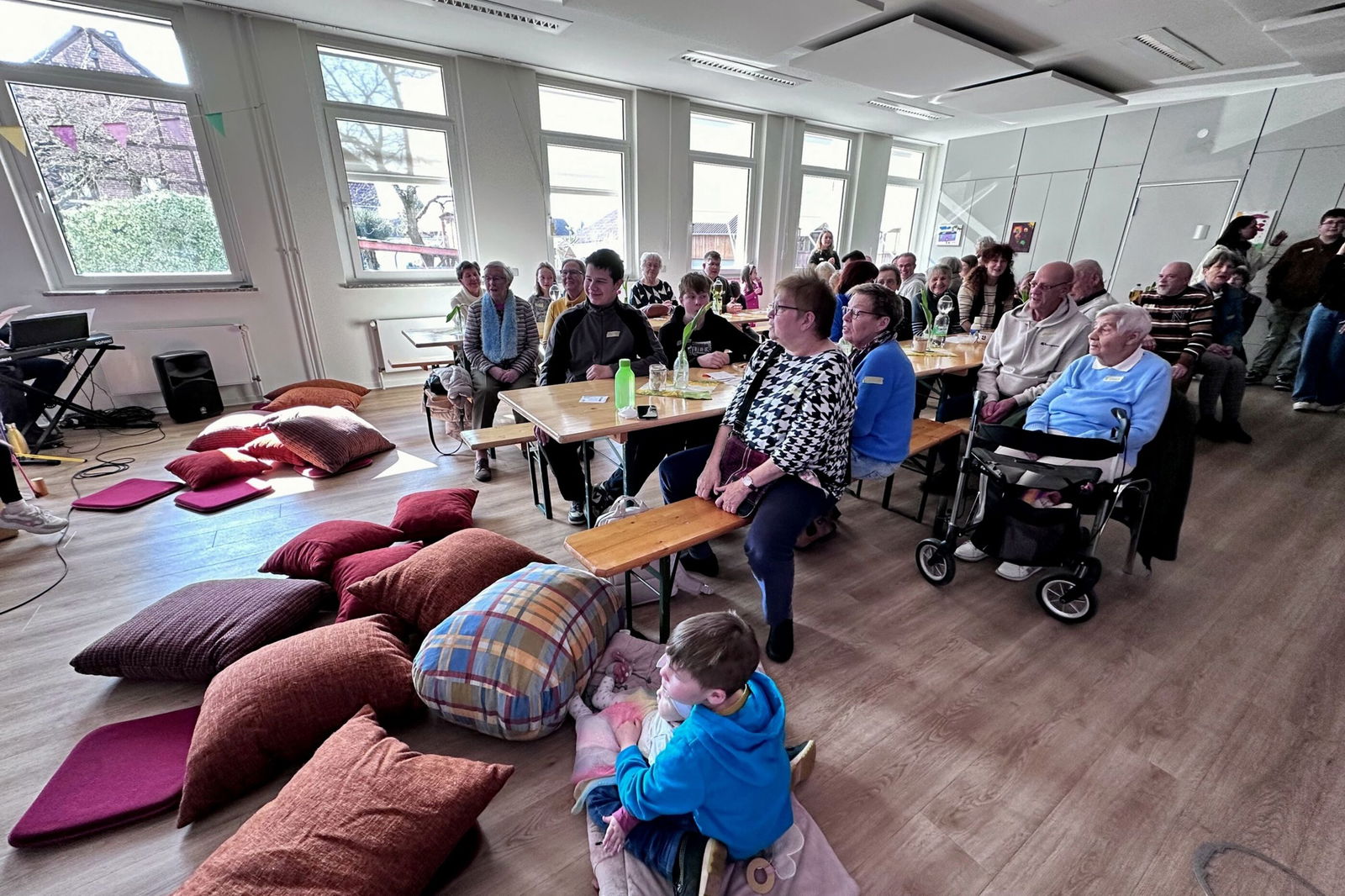 Mehrere Menschen sitzen an Tischen in einem Raum. Links sitzen Kinder auf großen, bunten Kissen.