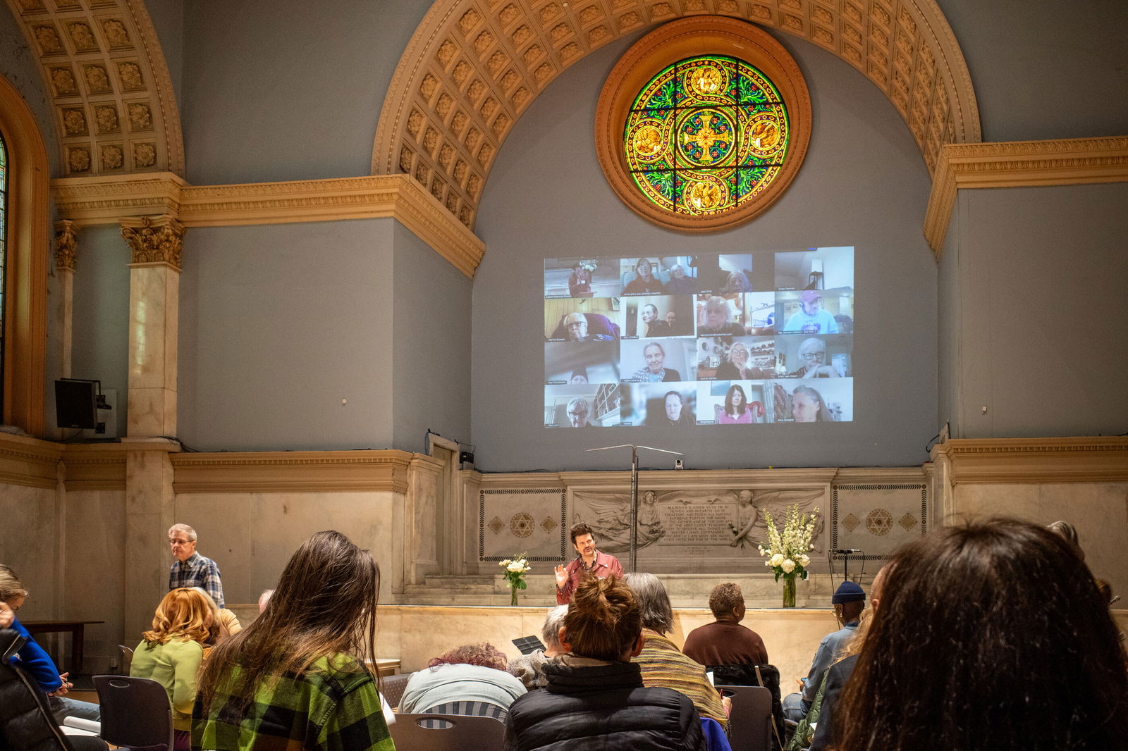 Blick in eine Kirche mit großem gelben Buntglasfenster. Unter diesen ist eine Videokonferenz mit etwa 25 Teilnehmenden projiziert. Davor sitzen Menschen.