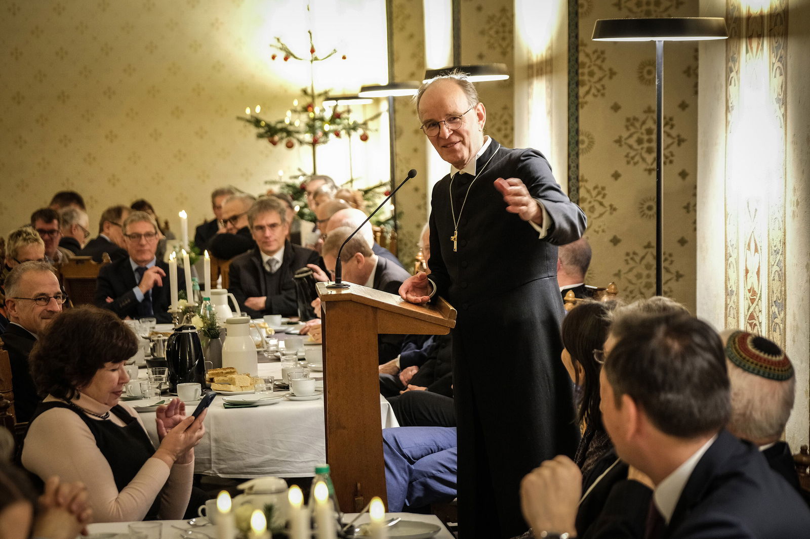 Mehrere Menschen sitzen an gedeckten Tischen, eine Person steht und spricht.