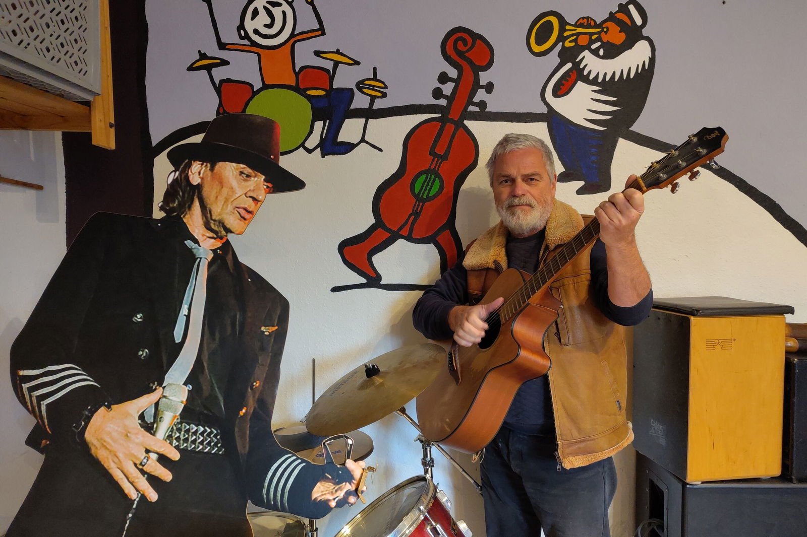 Zwei Personen vor einer bunten Wand, eine spielt Gitarre, der andere ist aus Pappe und sieht Udo Lindenberg ähnlich.