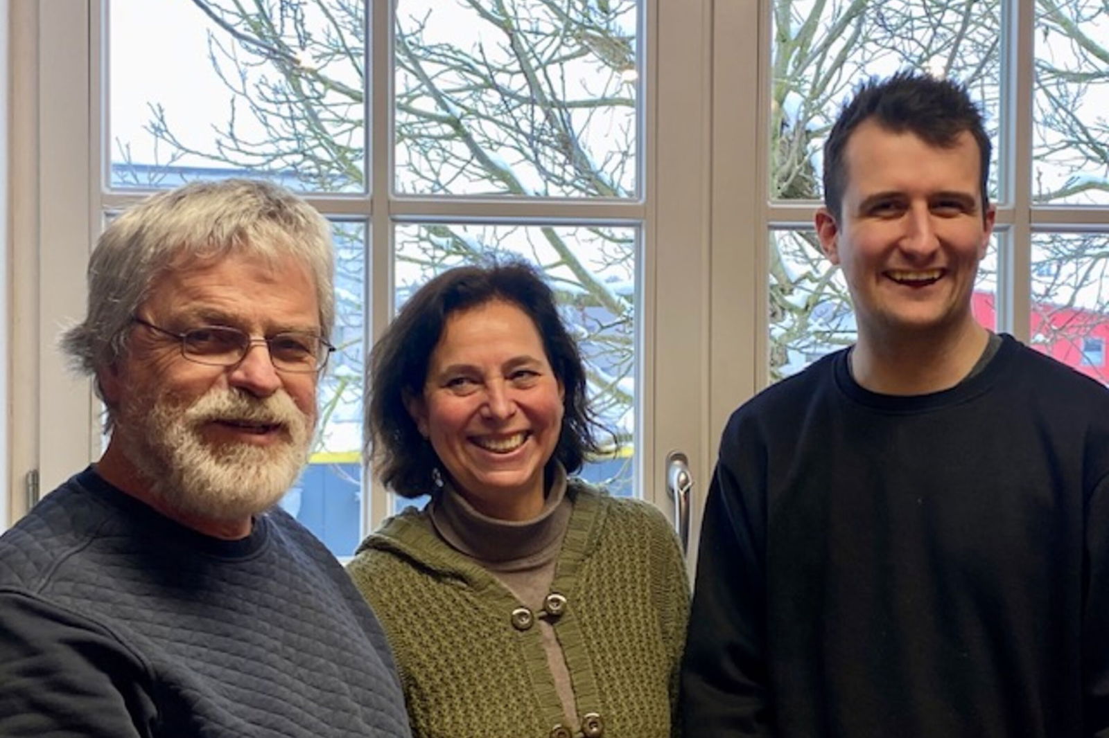 Zwei männlich und eine weiblich gelesene Person stehen vor einem Fenster.