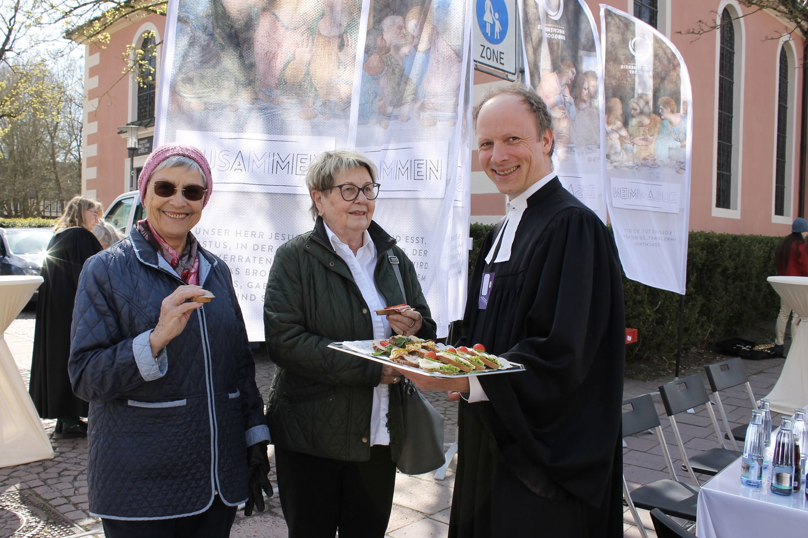 Drei Personen stehen vor einer Kirche, hinter ihnen wehen Banner mit einer Abendmahlsszene.