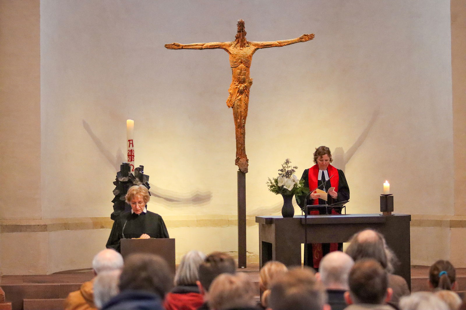 Zwei weiblich gelesene Personen in einer Kirche vor einem Publikum, mit einem großen Kruzifix im Hintergrund.