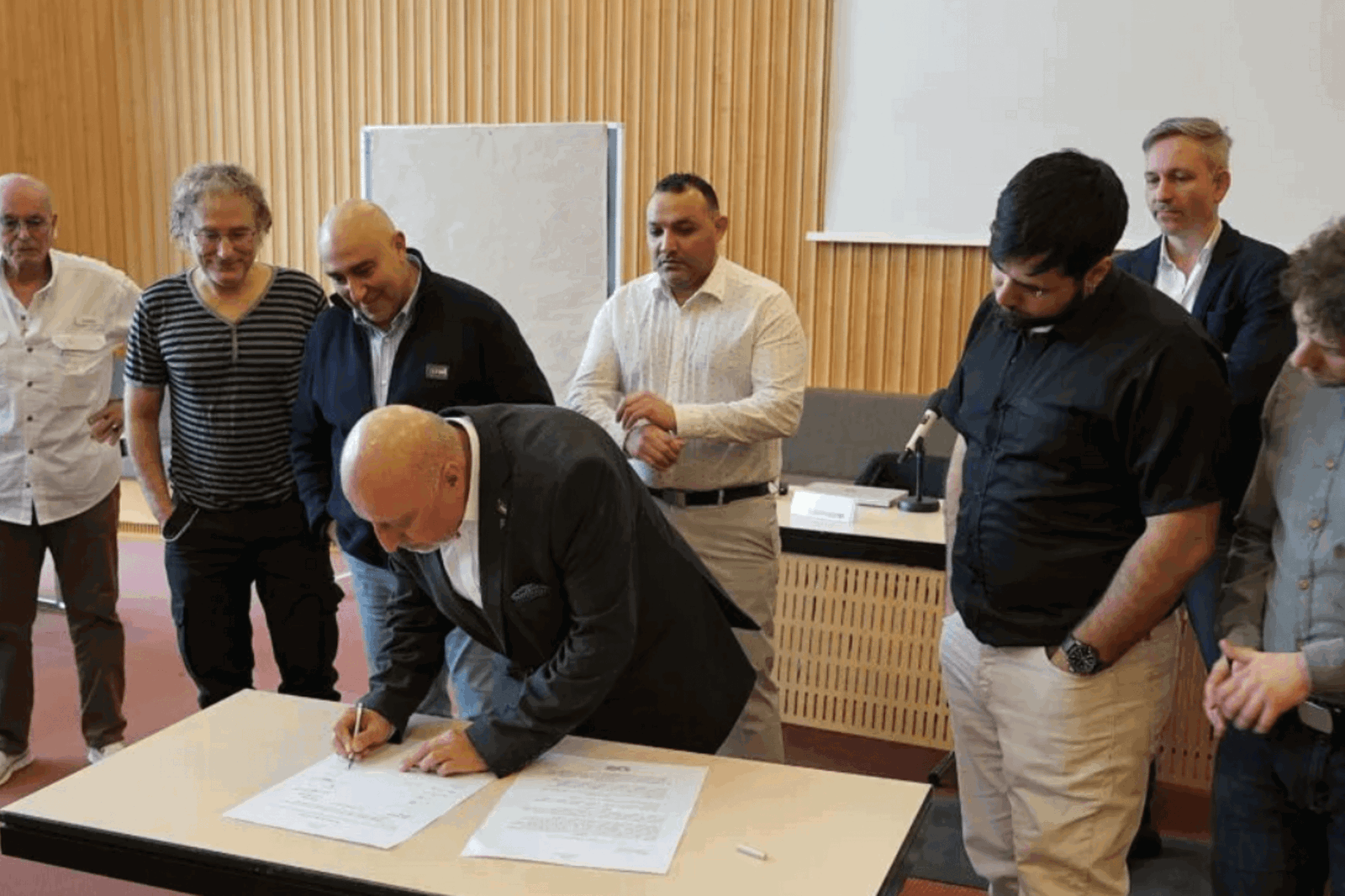 Eine männlich gelesene Person unterschreibt etwas, sieben andere Personen stehen um sie herum.