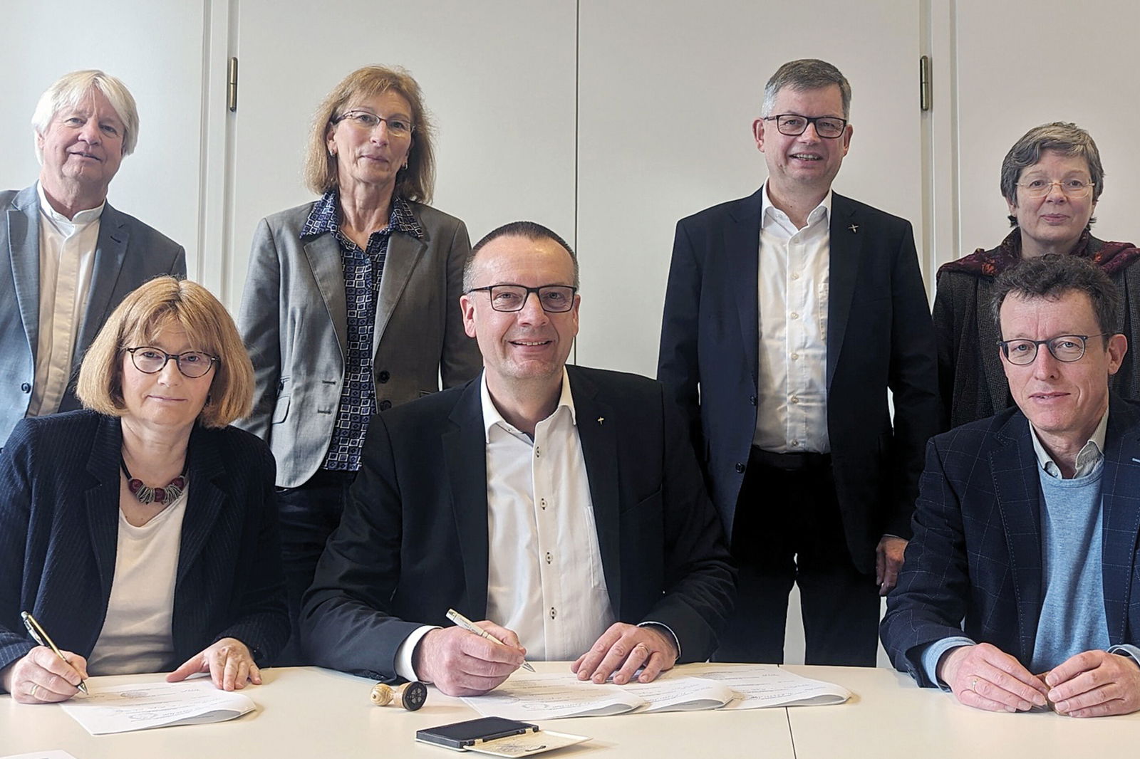 Drei weiblich und vier männlich zu lesende Personen stehen und sitzen hinter einem Tisch. Zwei von ihnen unterschreiben Unterlagen.