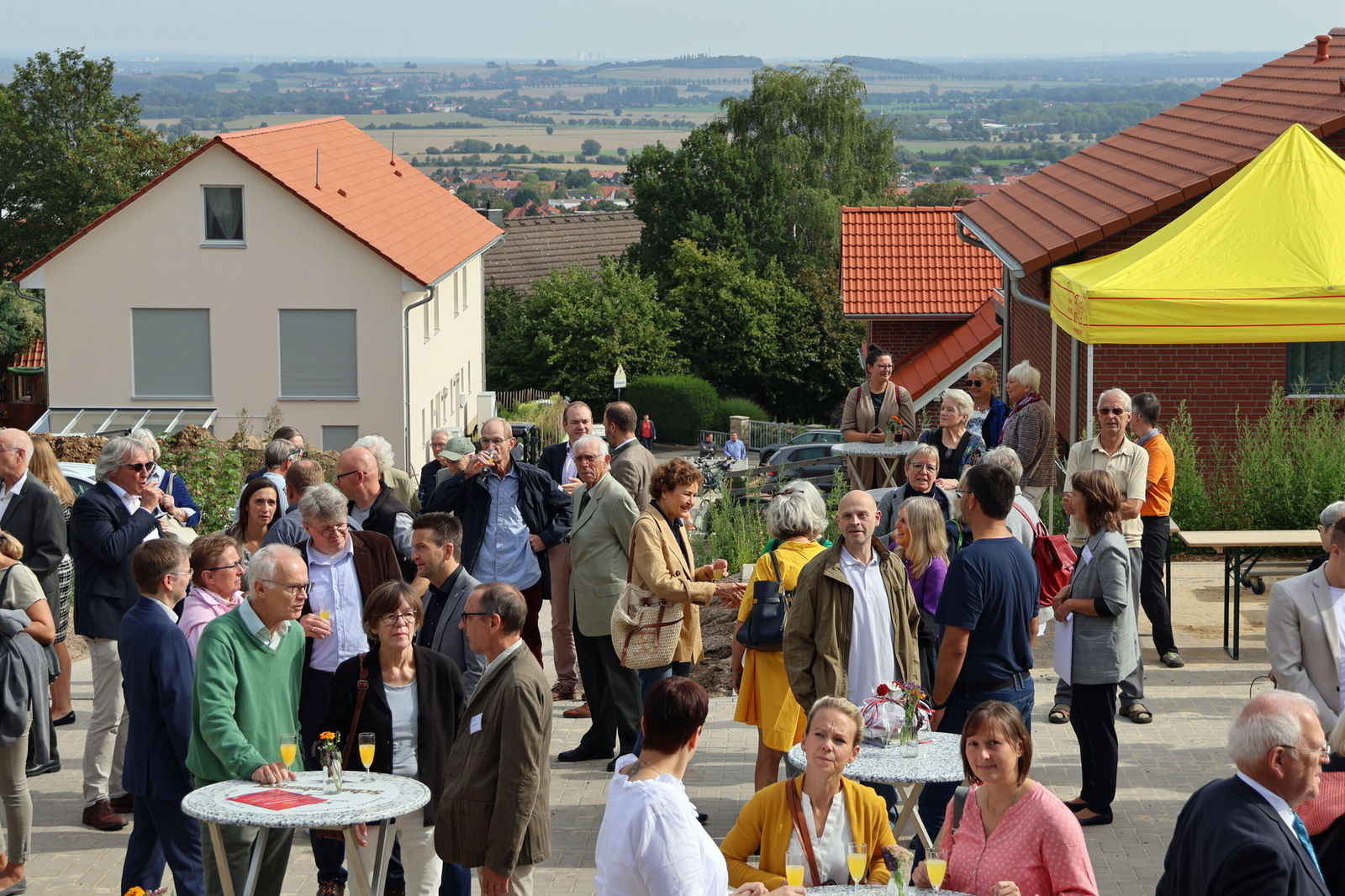 Mehrere Menschen stehen in Grüppchen zusammen an Stehtischen. Im Hintergrund öffnet sich der Blick in weites, flaches Land.