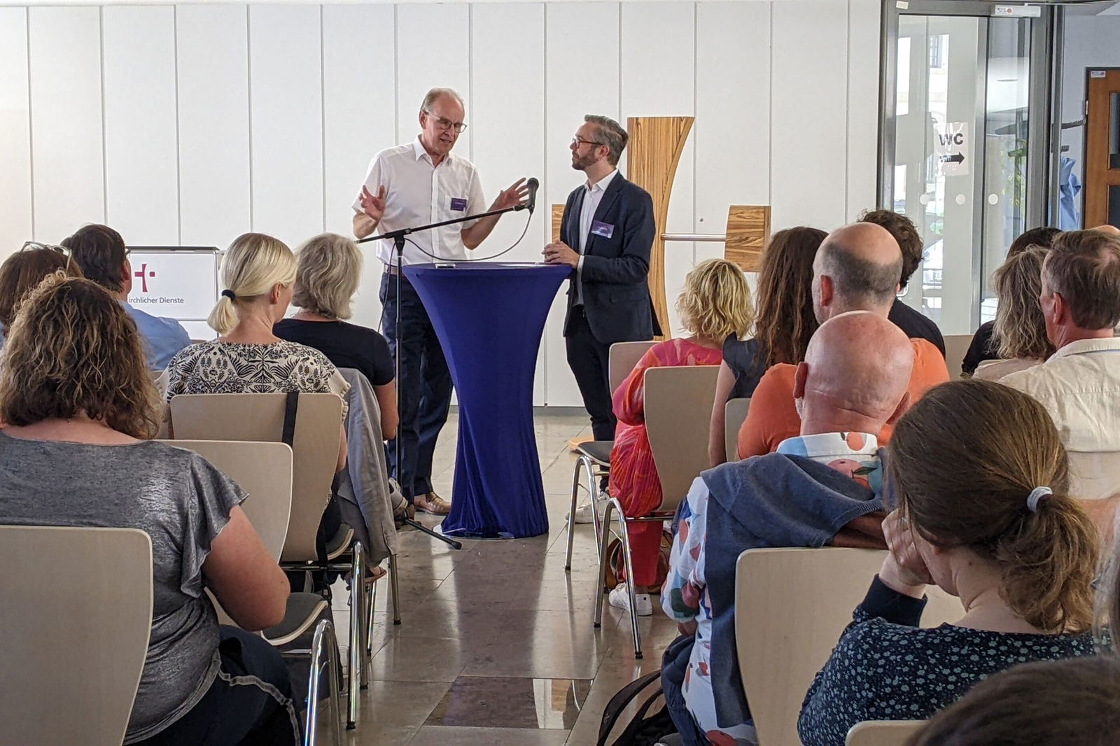 Zwei Männer, einer in weißem Hemd, einer in dunklem Anzug, stehen an einem Stehtisch vor mehreren sitzenden Menschen. 