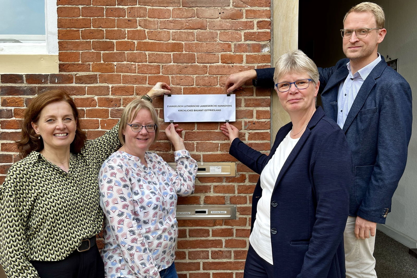 Vier Personen stehen vor einer Eingangstür und legen Hand an Klammern, die ein provisorisches Schild aus Papier festhalten.