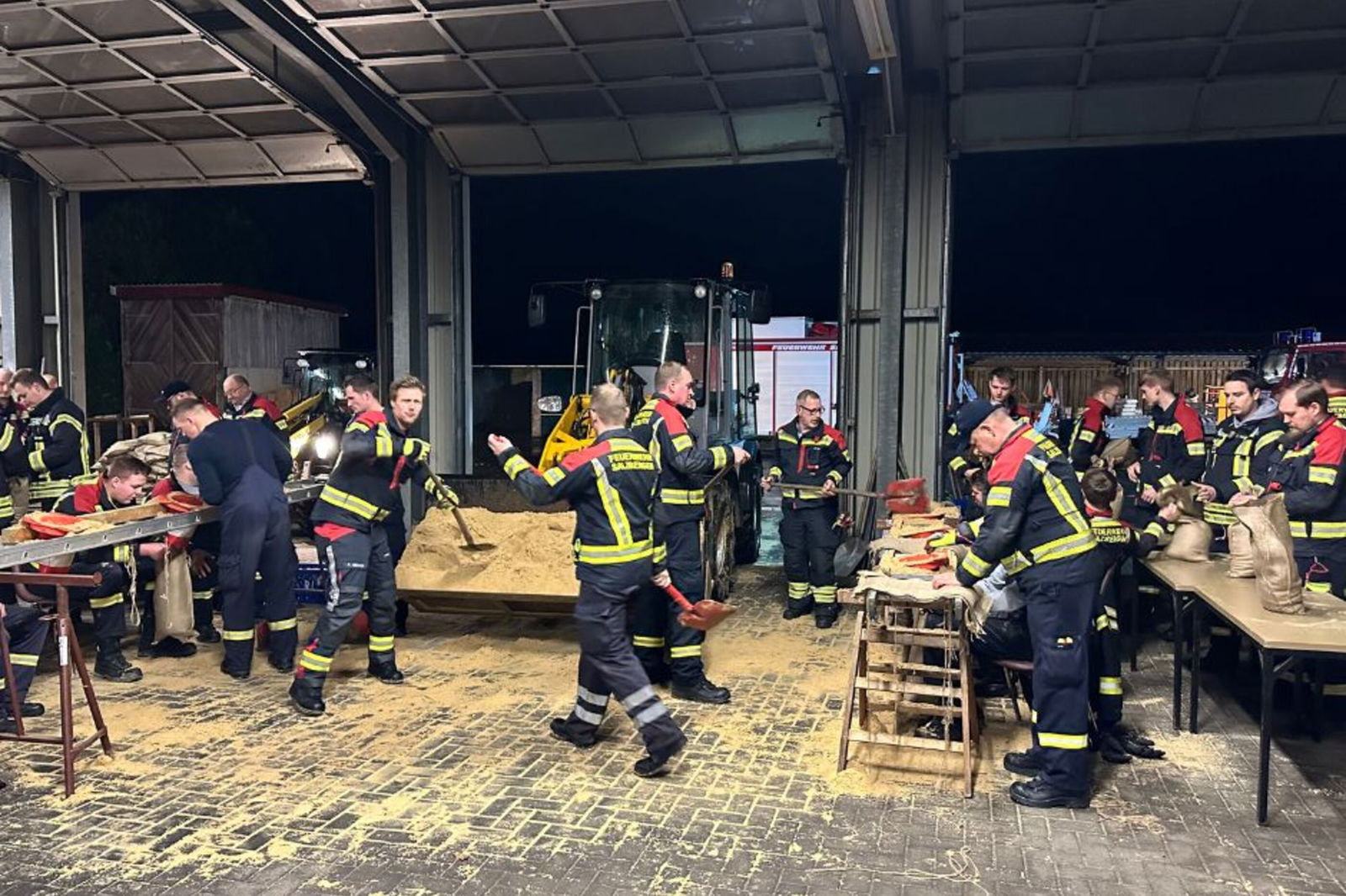 Etwa zwei Dutzend Feuerwehrmänner stehen an Tischen oder neben einem Container voll Sand und füllen Sandsäcke.