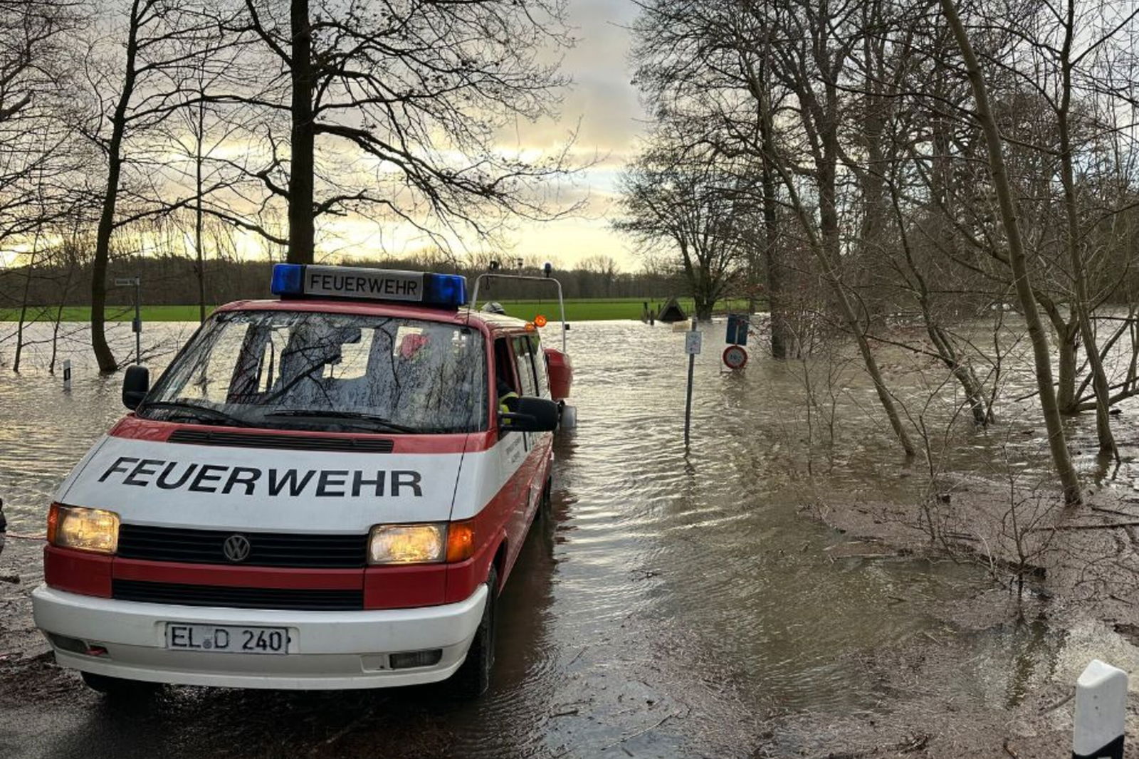 Ein Einsatzwagen einer Feuerwehr steht im Wasser, das einen Weg über- und Bäume umspült.