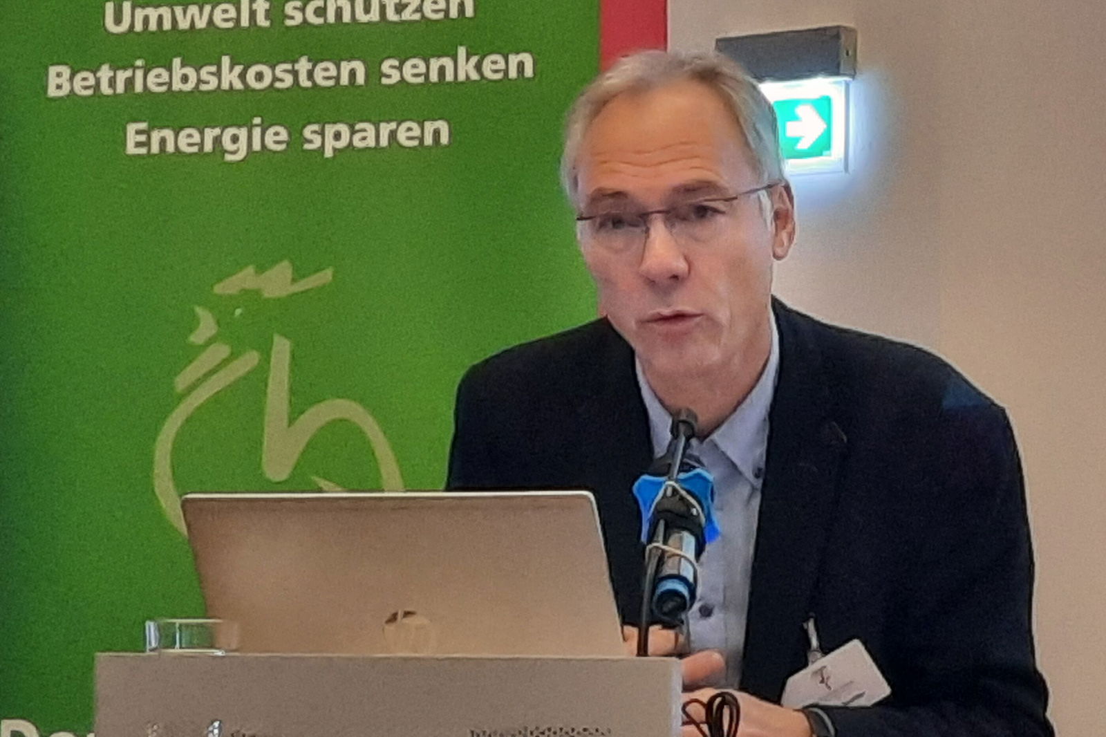 Ein Mann mit kurzen grauen Haaren und Brille, dunklem Jackett und blauem Hemd steht an einem Redepult. Im Hintergrund ein Banner: weiß auf grün steht dort: Umwelt schützen - Betriebskosten senken - Energie sparen.