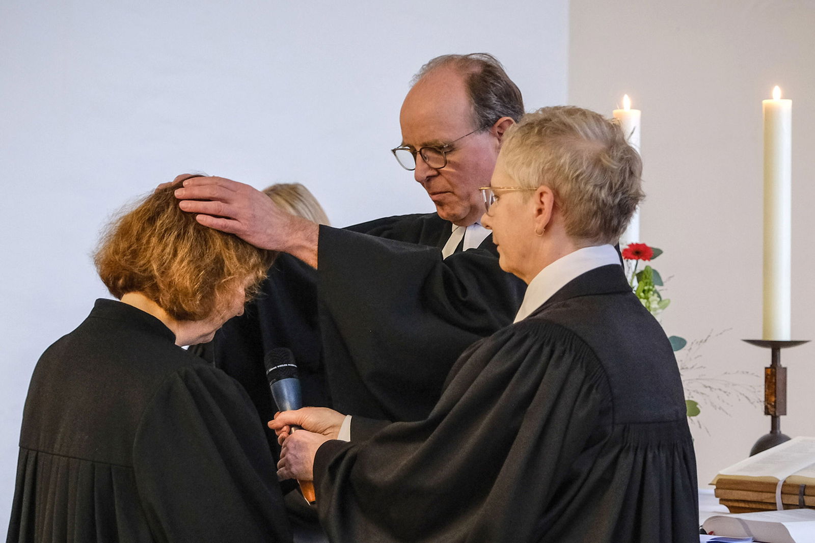 Ein Mann legt seine Hände auf den Kopf einer blonden Frau. Daneben steht eine Frau mit kurzen, grauen Haaren. Alle tragen Talare. Im Hintergrund stehen Kerzen auf einem Altar.