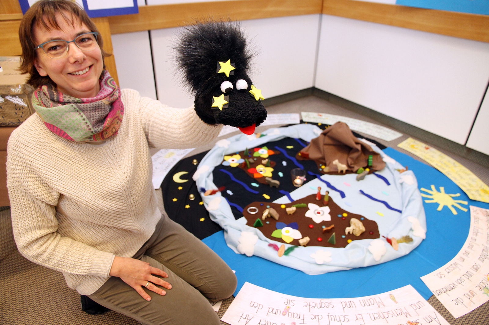 Eine Frau hält eine Handpuppe. Hinter ihr liegt ein Bodenbild zur Schöpfung.