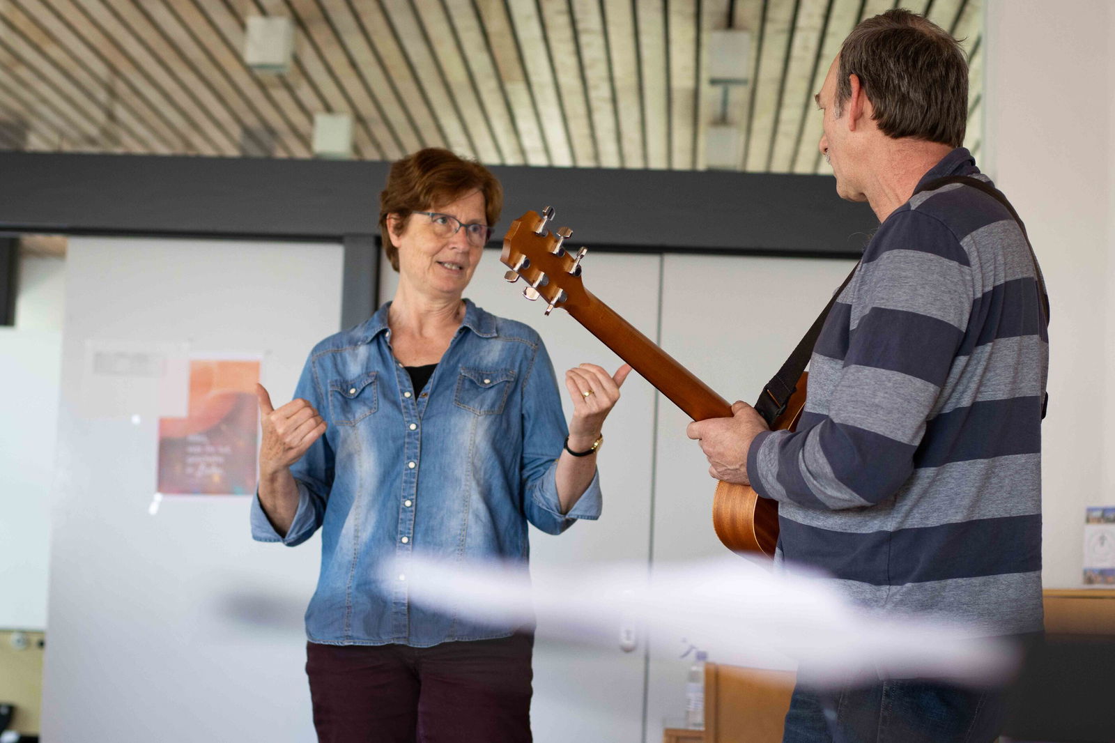 Eine weiblich gelesene Person in Jeanshemd steht neben einer männlich gelesenen Person in gestreiftem Shirt, der eine Gitarre um den Hals hat.
