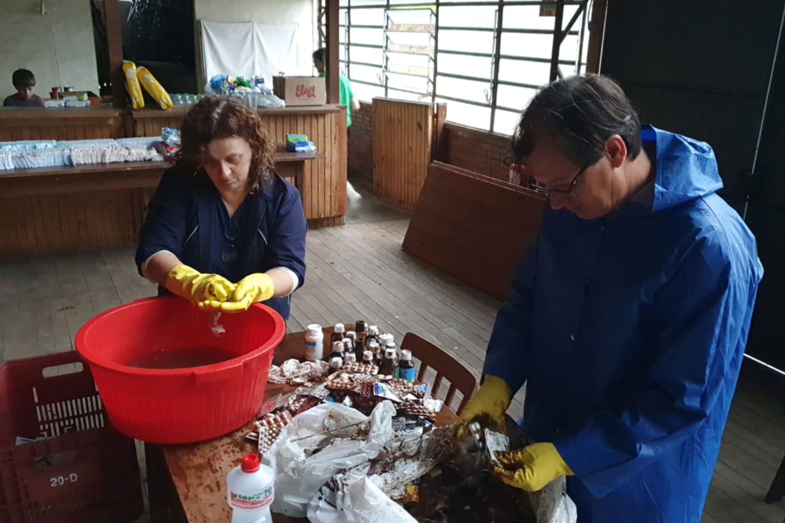 Ein Raum mit Tischen. Im Vordergrund ein Tisch auf dem verschiedenen Dinge liegen. An dem Tisch stehen zwei Personen. Sie tragen gelbe Handschueh und reinigen Gegenstände.