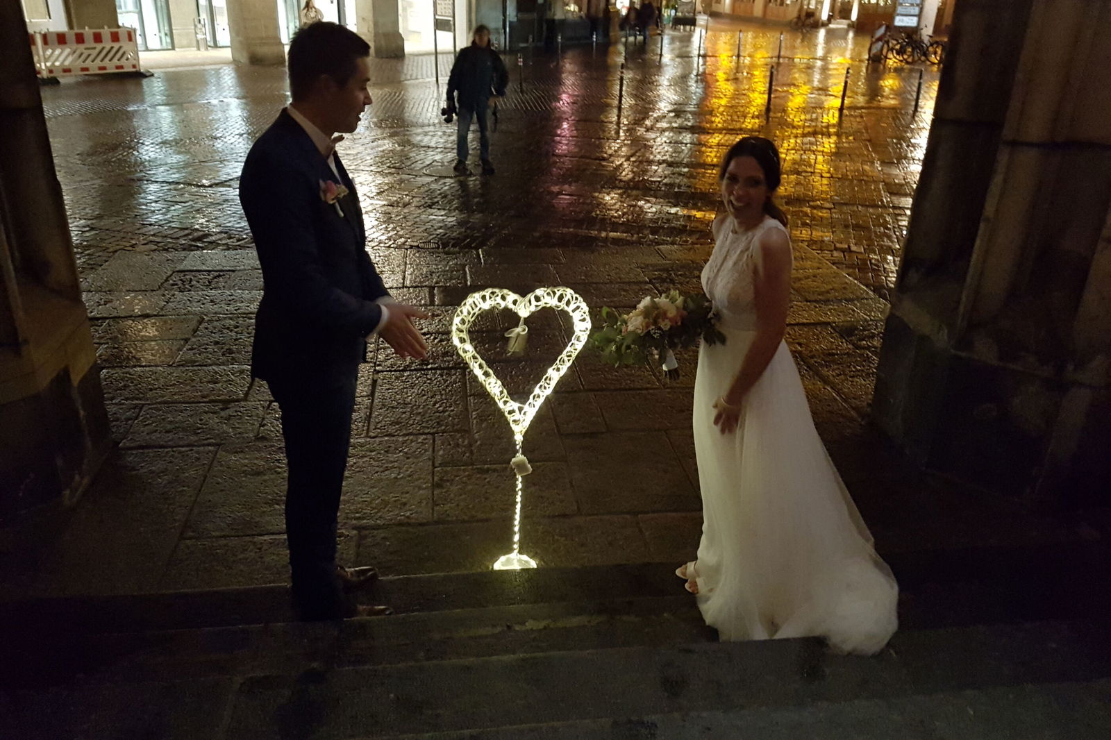 Eine männlich gelesene Person im Anzug und eine weiblich gelesene in einem Brautkleid stehen links und rechts eines kerzenerleuchteten Herzens auf einer spiegelnassen Straße.