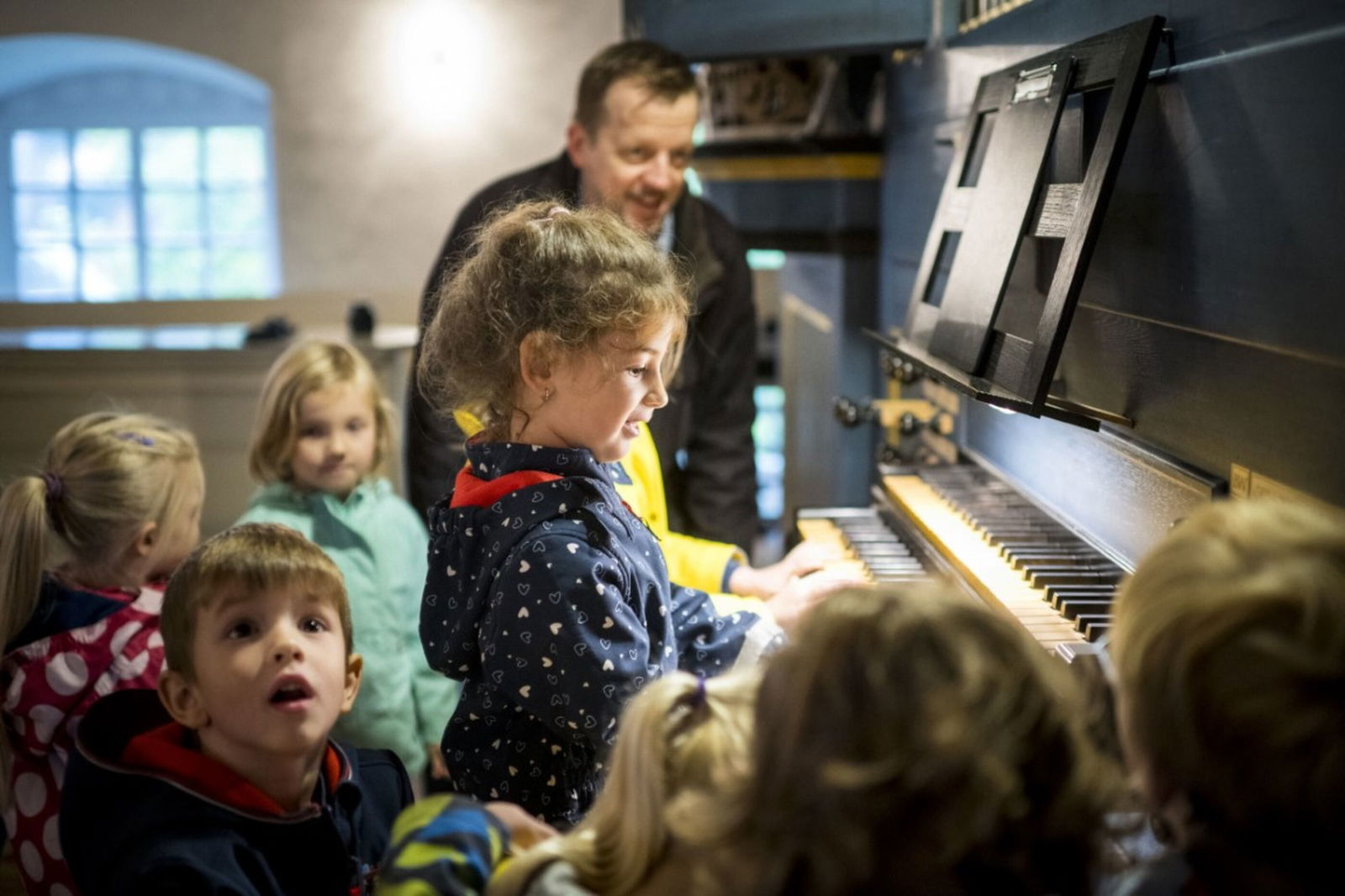 Mehrere Kinder haben sich um eine Orgel versammelt, ein Kind spielt darauf. Im Hintergrund verschwommen eine männlich gelesene Person, die zuschaut.
