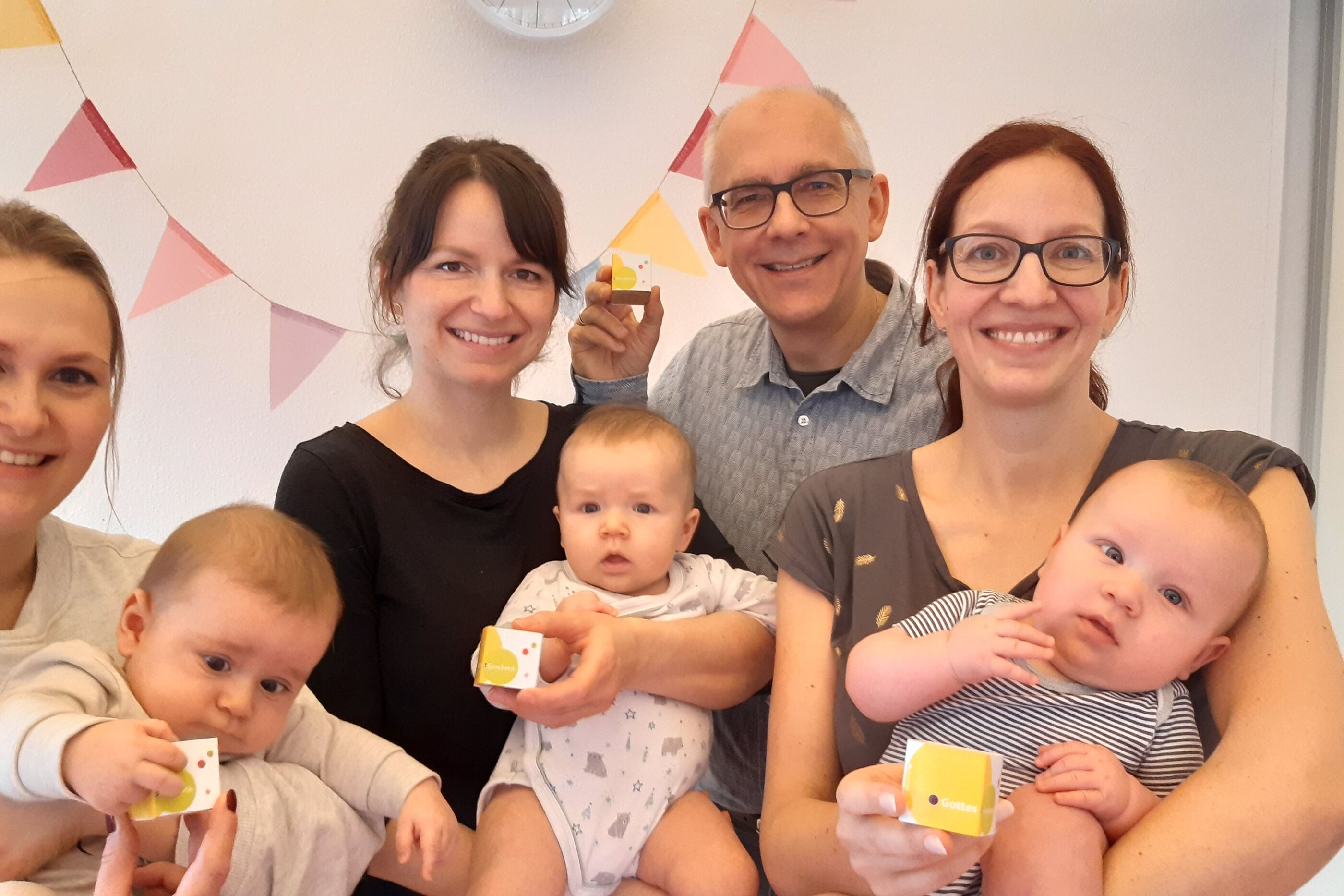 eine männlich gelesene Person und weiblich gelesenen Personen mit Babys halten kleine Würfel aus Pappe in der Hand.