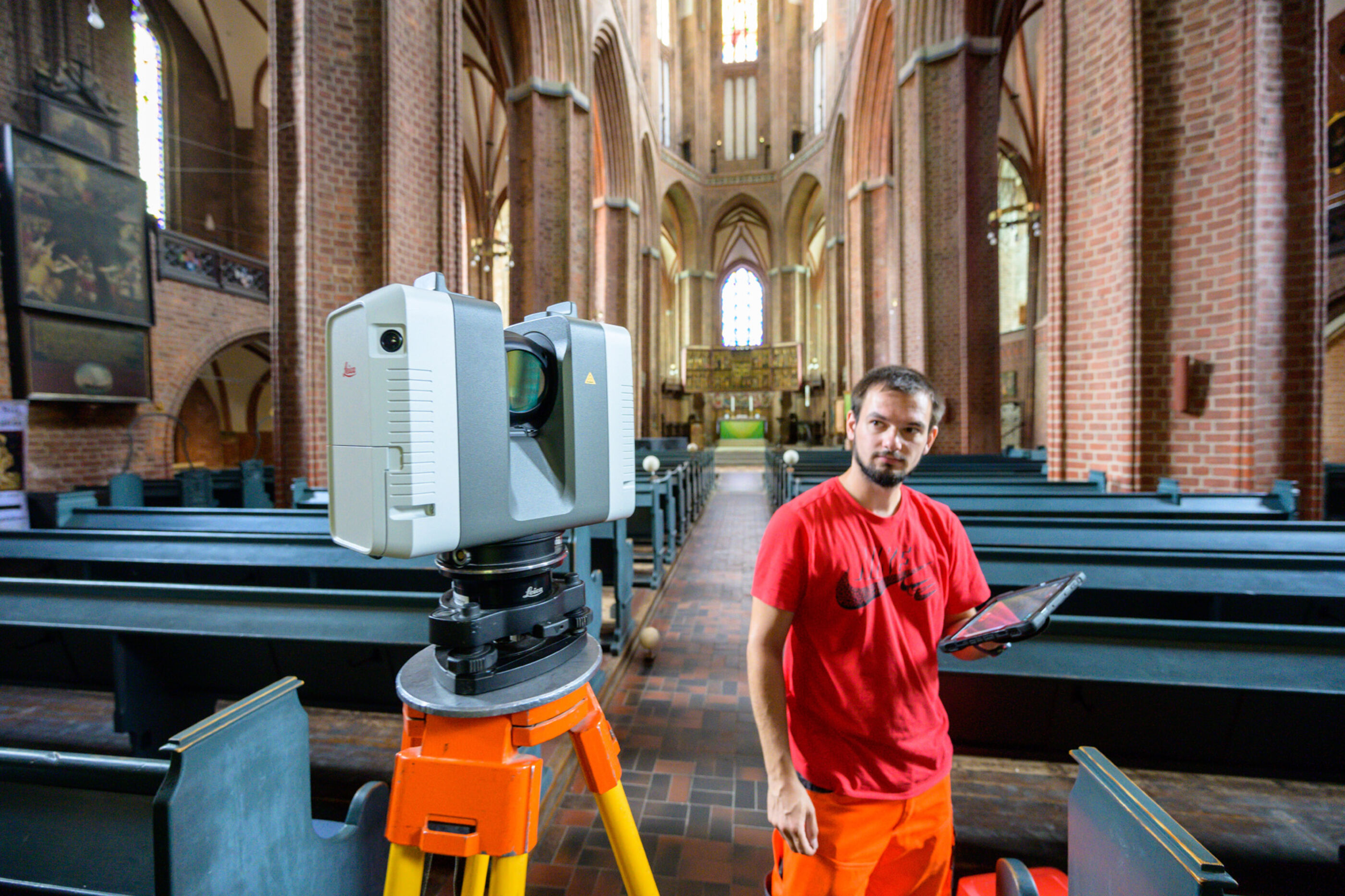 Im Mittelgang steht zwischen den Kirchenbänken ein Laserscanner mit Kamera. Daneben steht ein Mann mit roten Klamotten.