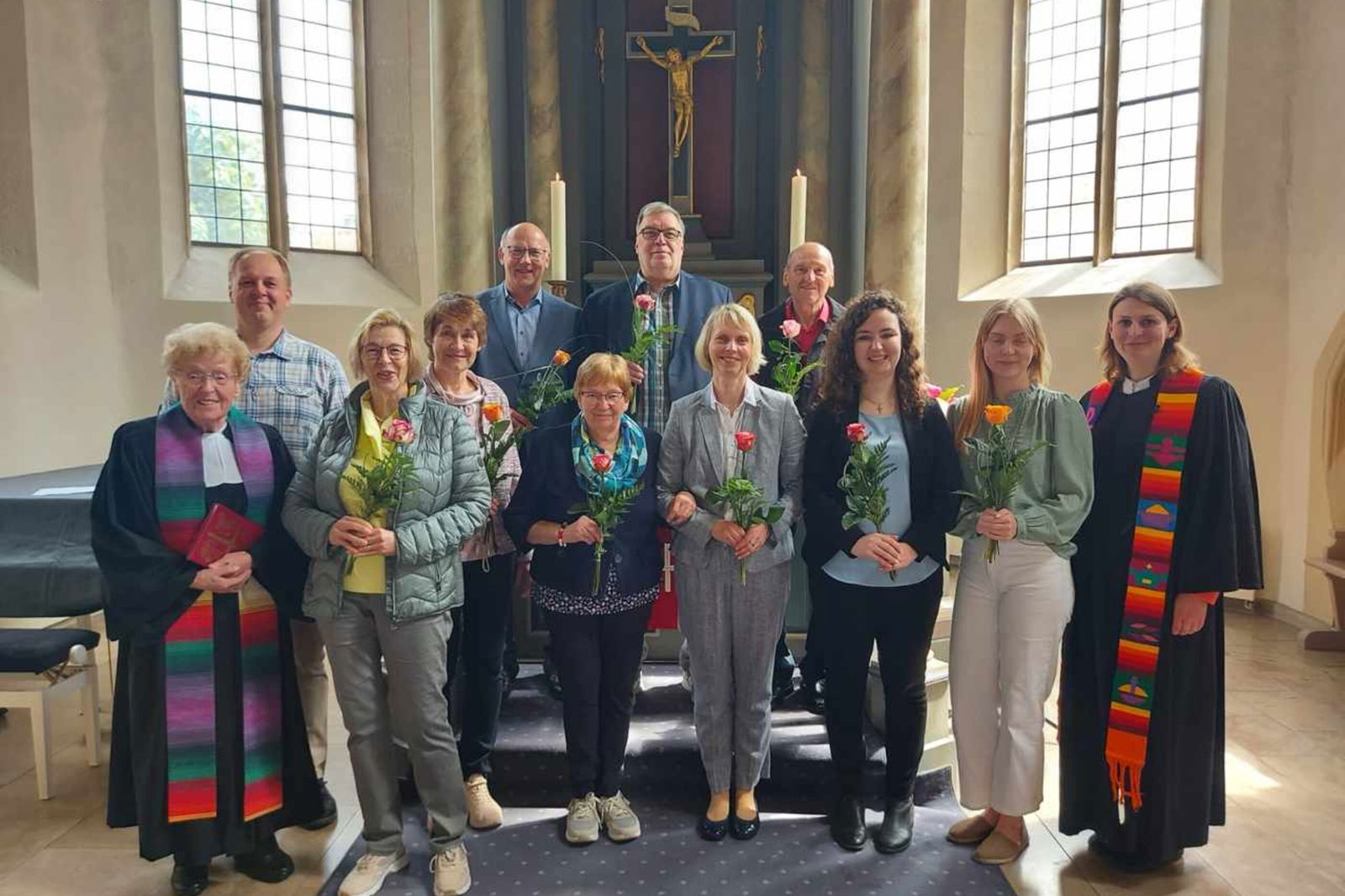 Mehrere Menschen stehen im Altarraum einer Kirche, sie halten Blumen in den Händen. Links und rechts stehen zwei Personen in Talaren.
