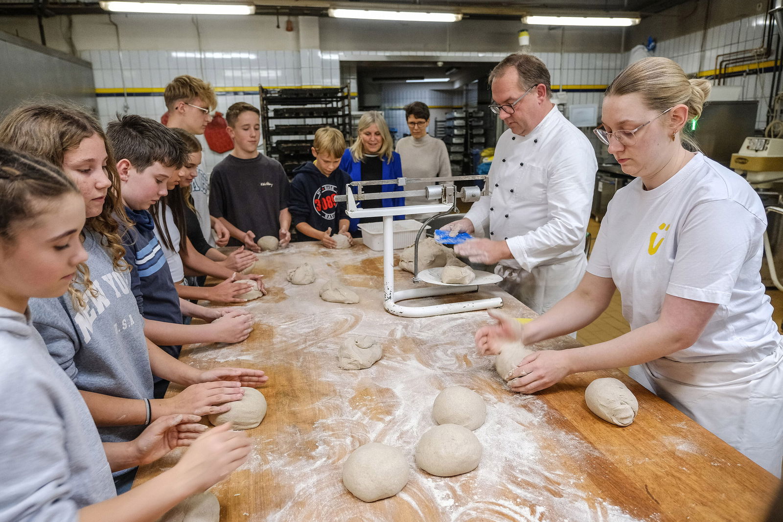 Mehrere junge Menschen und zwei Erwachsene in weißen Kitteln stehen an einem Tisch und kneten Teige, Mehl auf dem Tisch verteilt.