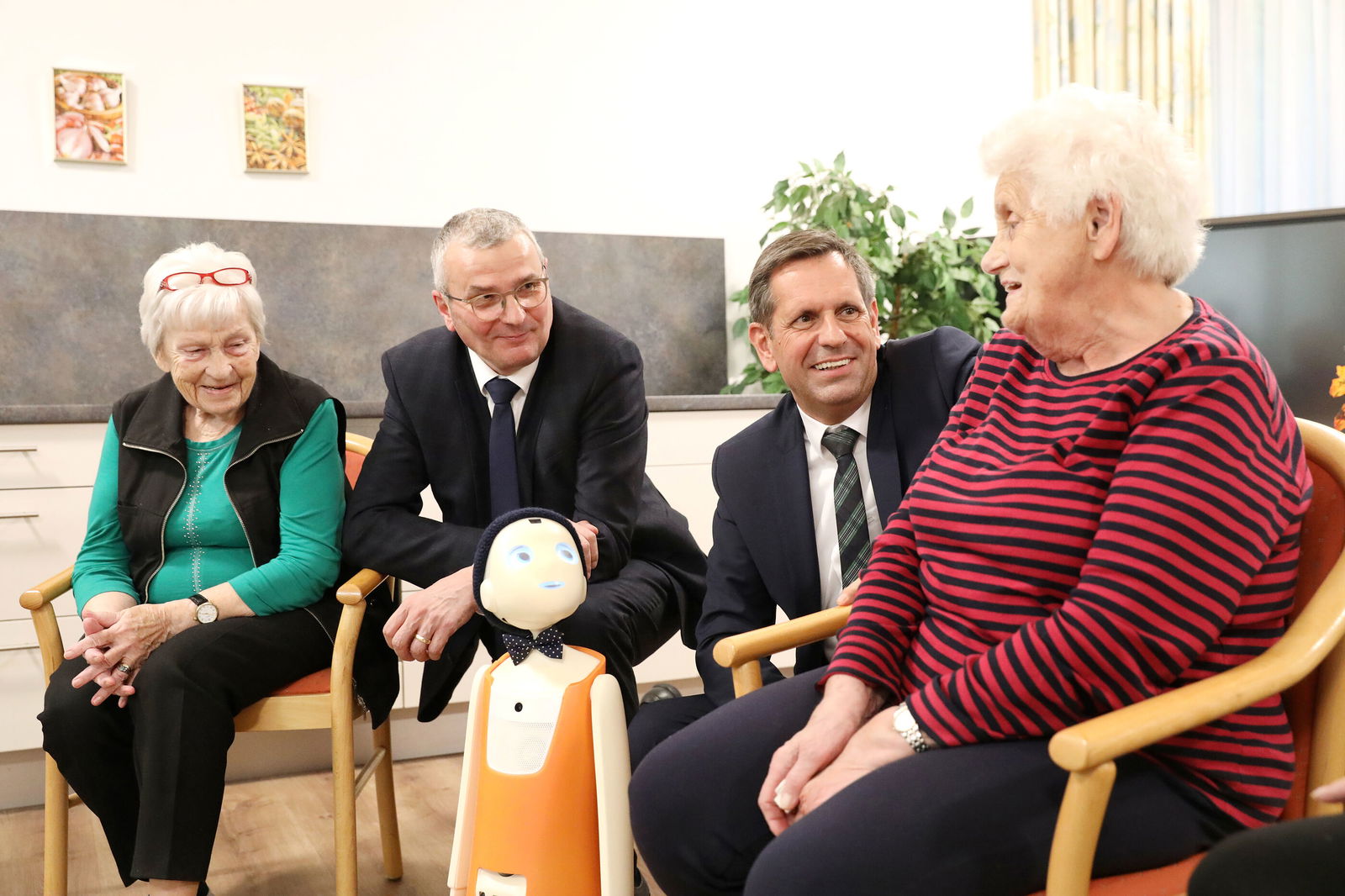 Vier Personen sitzen bzw. knien nebeneinander, vor ihnen ein weiß-orangener Roboter mit Gesicht.