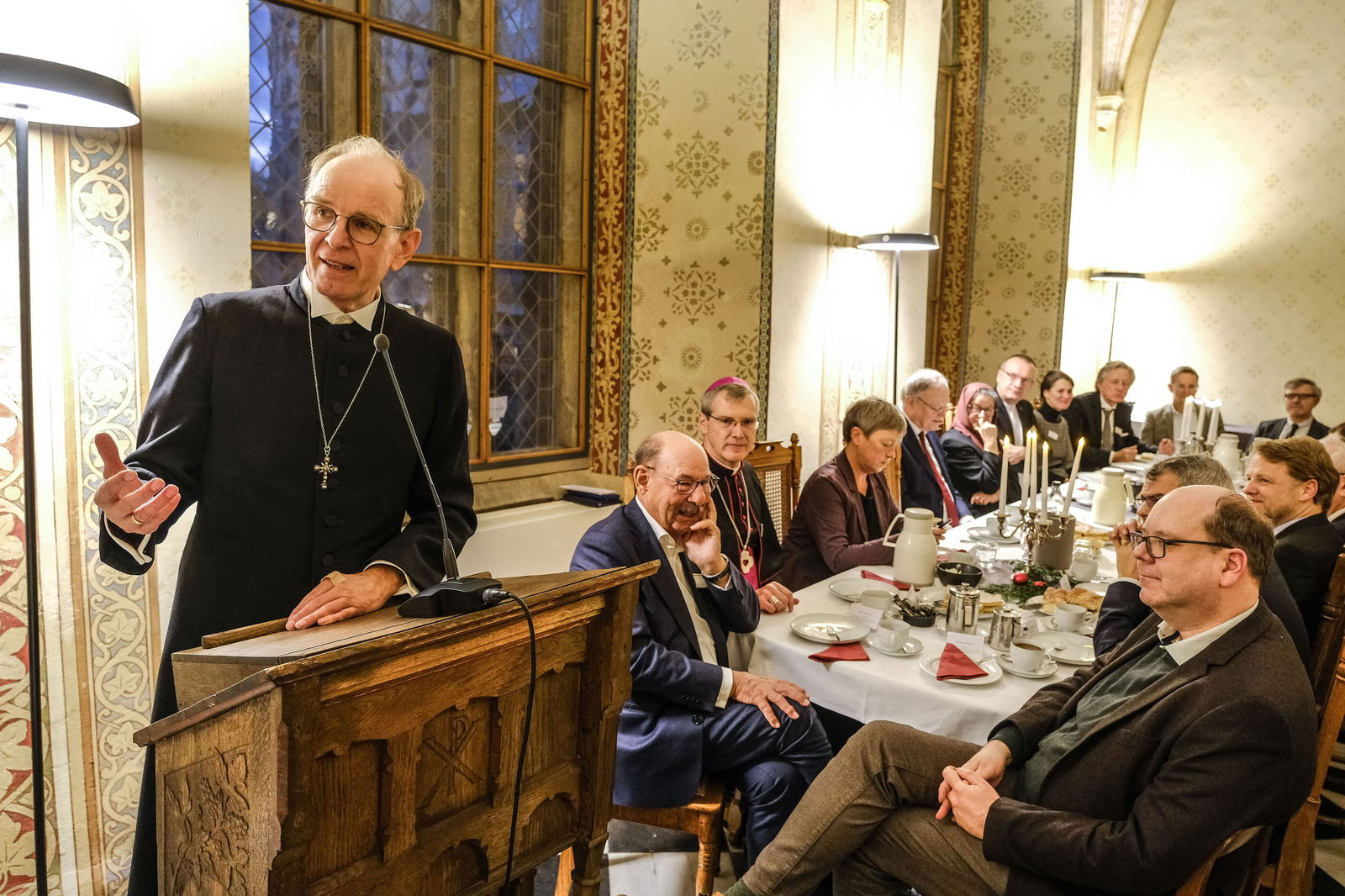 Eine männlich zu lesende Person steht an einem Pult und spricht zu eine Gruppe von Personen, die an einem gedeckten Tisch sitzen.