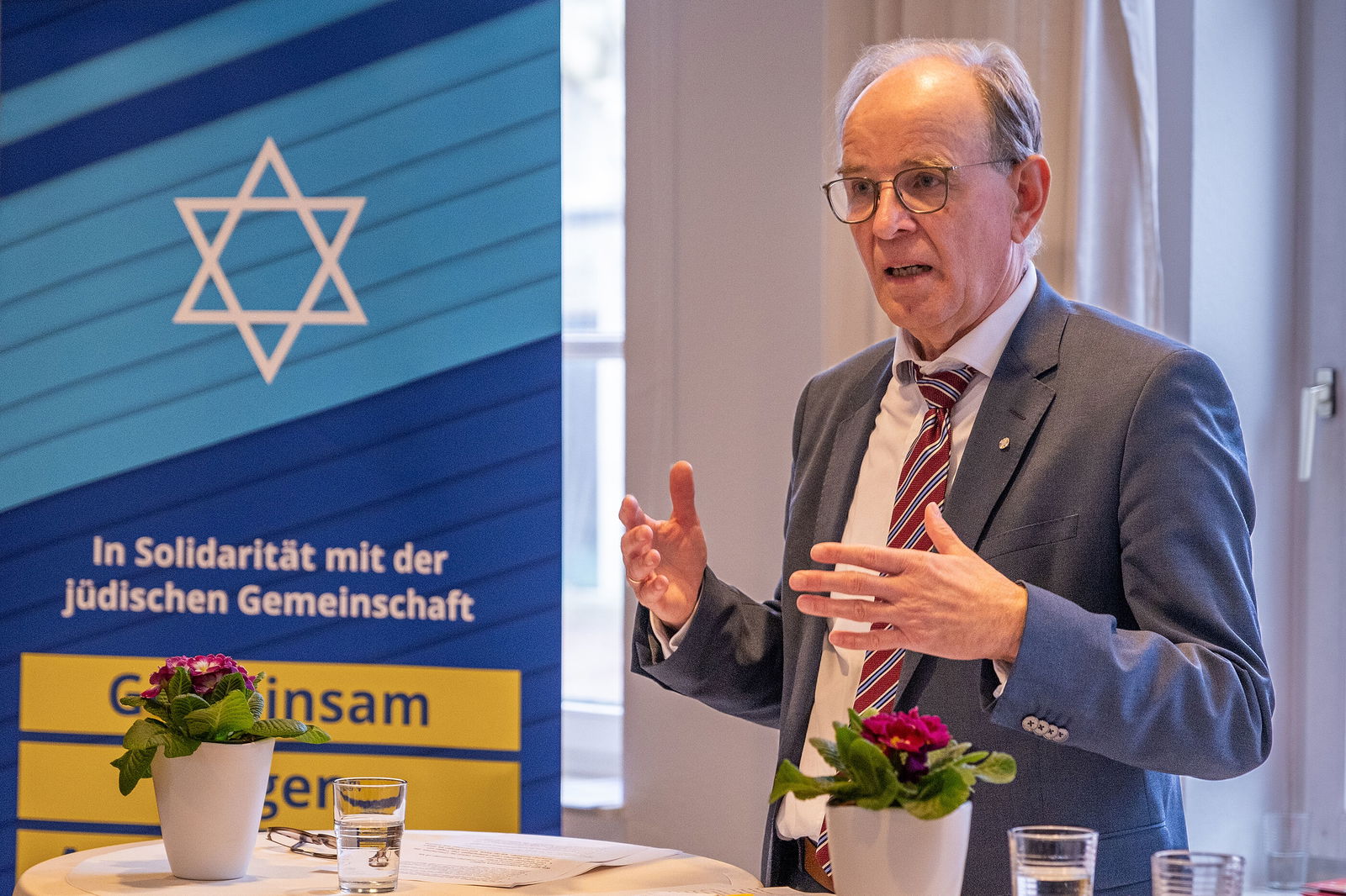 Eine männlich gelesene, ältere Person mit Brille und kurzen grauen Haaren in Anzug und Krawatte, steht an einem Stehtisch und gestikuliert. Im Hintergrund ein Banner, auf dem ein weißer Davidstern auf blauem Untergrund zu sehen ist.