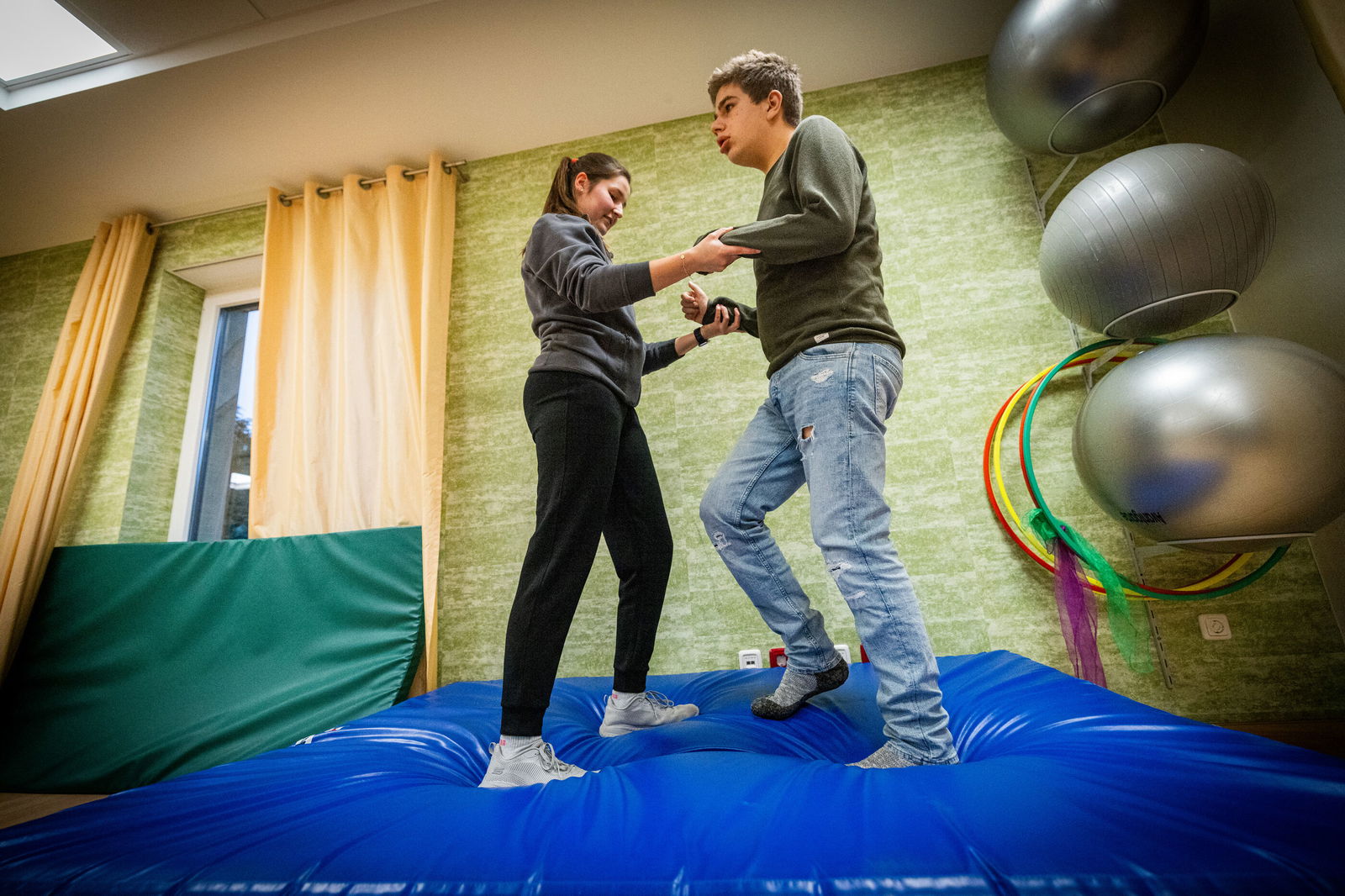 Eine ältere, weiblich gelesene Person hält eine jüngere, männlich gelesene an den Händen, während sie auf einer weichen Matte balancieren.