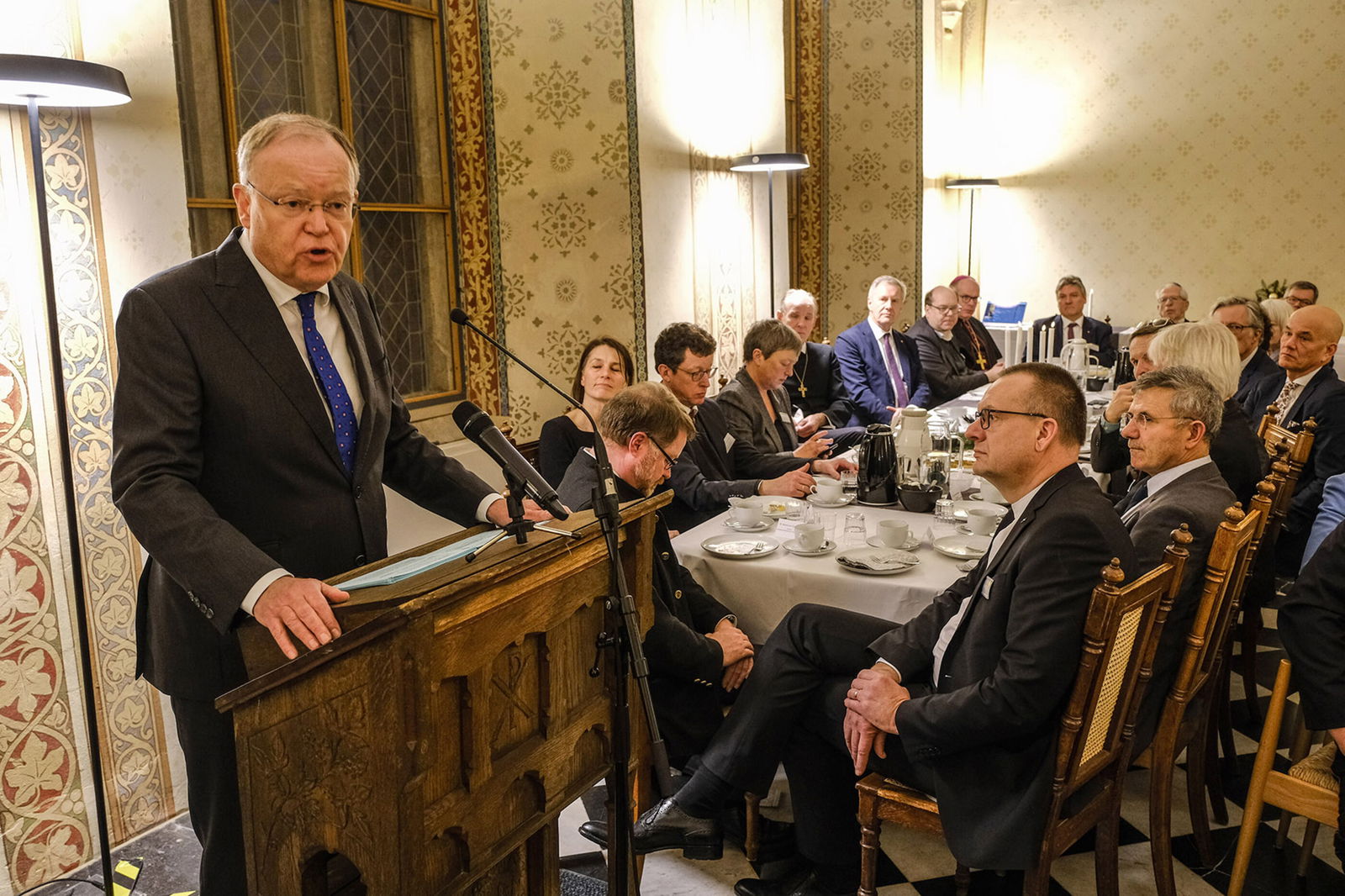Ein Mann im Anzug mit blauer Krawatte steht an einem Rednerpult. Links von ihm sitzen etwa ein Dutzend Menschen an einem gedeckten Tisch.