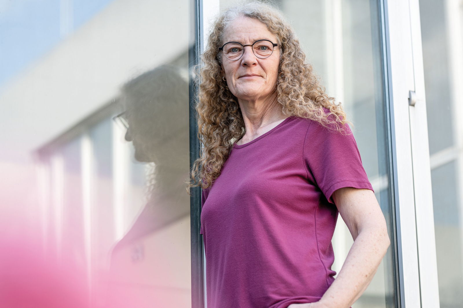 Eine weiblich gelesene Person in lila T-Shirt mit langen, lockigen Haaren steht an eine Wand gelehnt.