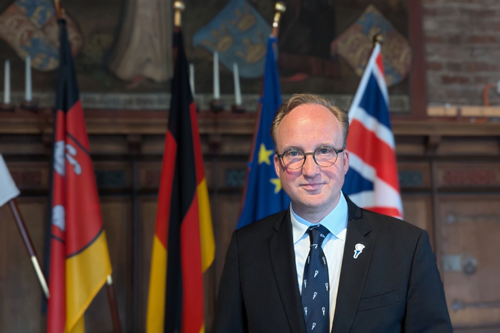 Eine männlich gelesene Person im Anzug vor deutschen, europäischer und britannischer Fahne.