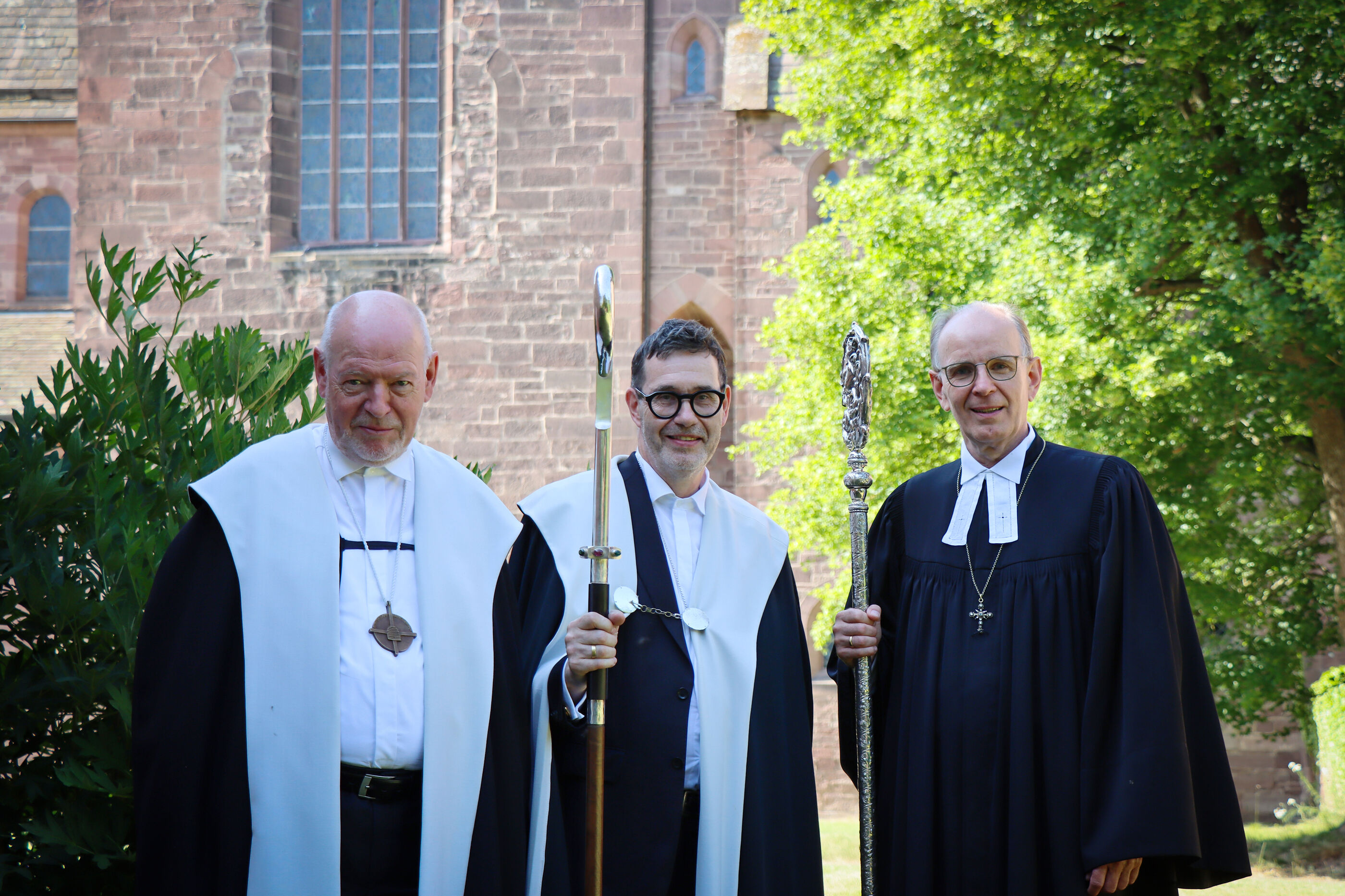 Drei Männer im Ornat vor einer Kirche.