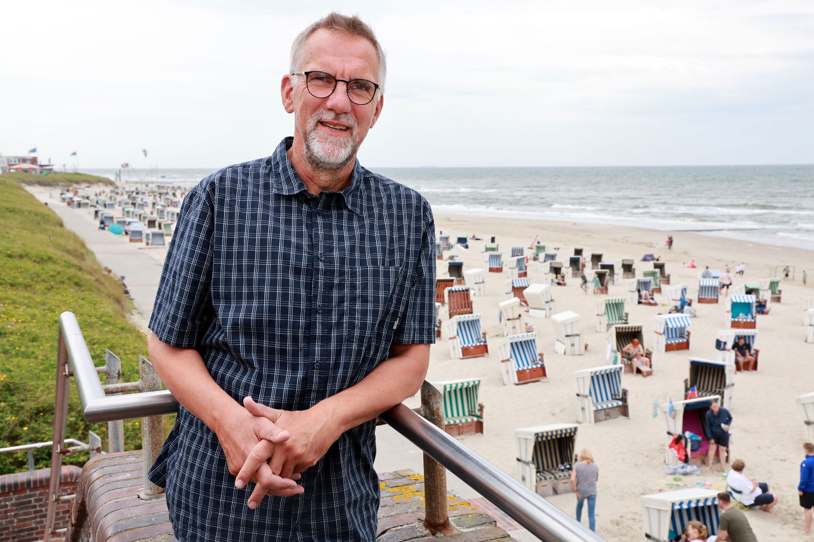 Eine männlich gelesene Person in kurzärmeligem Hemd an einer Balustrade, neben der ein Strand mit Strandkörben zu sehen ist.
