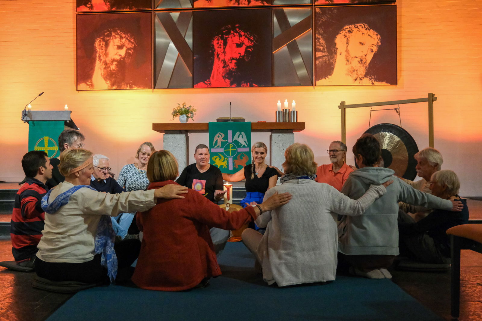Eine Gruppe von Menschen sitzt im Kreis, jeder Person legt der rechts von ihr sitzenden die Hand auf die Schulter.