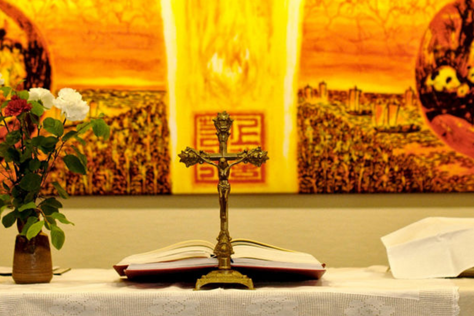 Vor einem leuchtend gelb-orangenen Hintergrund stehen ein Altarkreuz und Blumen.